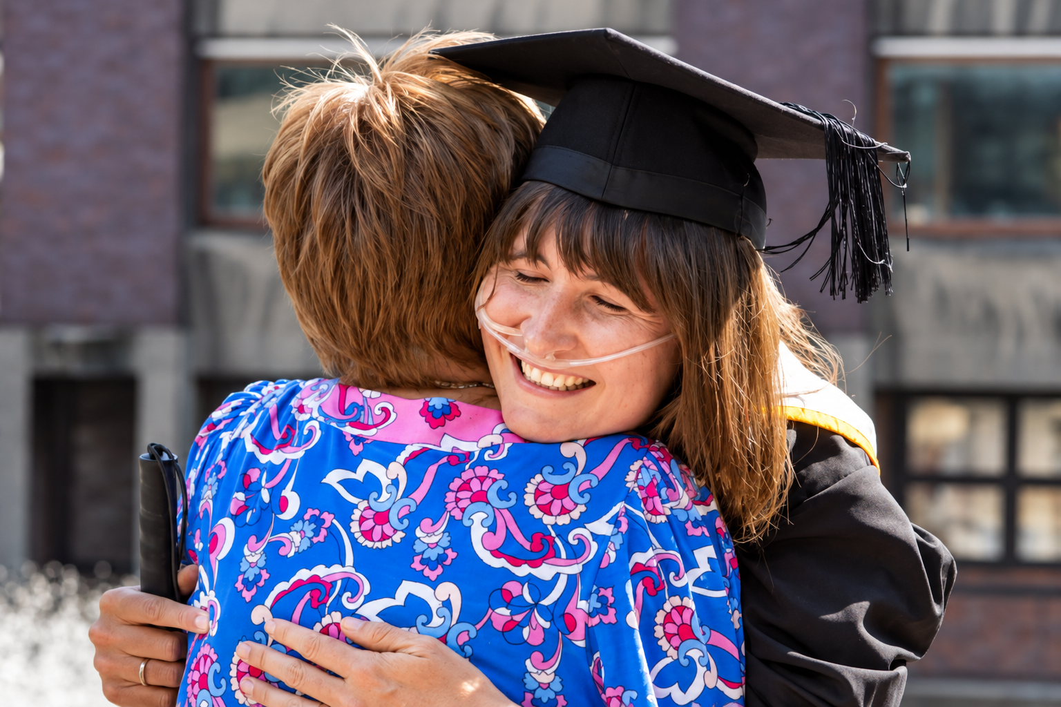 Alexandra wearing a black graduation cap and gown and a nasal oxygen cannula smiles while hugging another person outdoors after graduation.