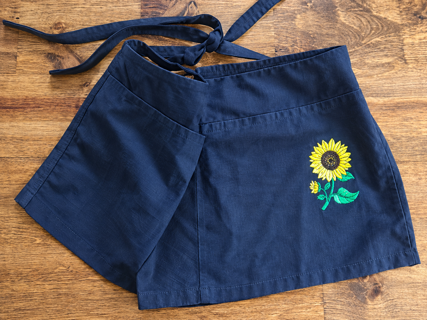 A navy-blue apron or wrap-style garment lies on a wooden surface, featuring an embroidered yellow sunflower with green leaves near the lower right side.