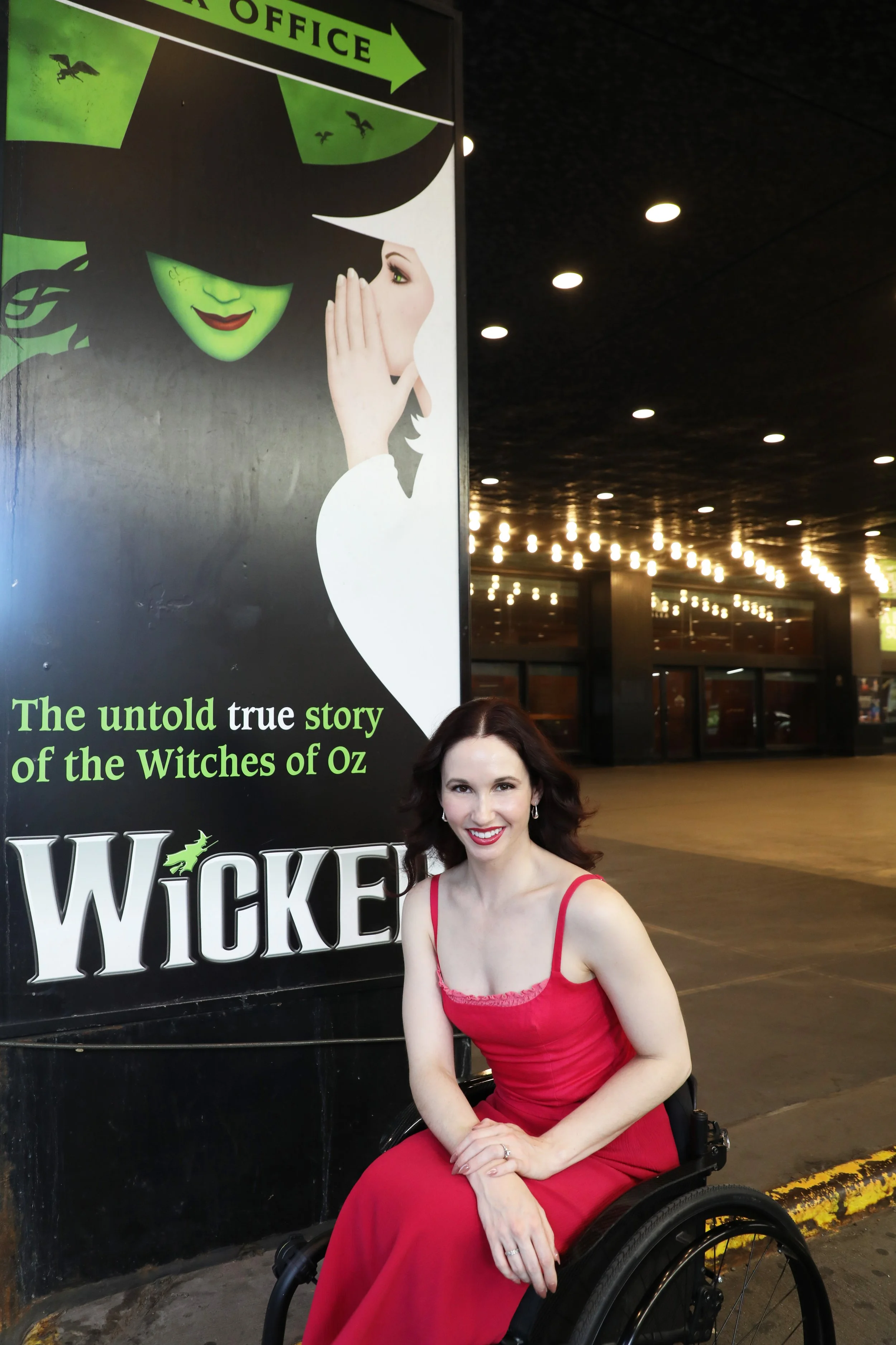 Jenna, wearing a dark pink summer dress, in front of the Wicked poster on Broadway