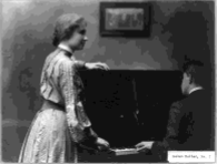 Helen Keller rests her arm on a standing piano to feel the music