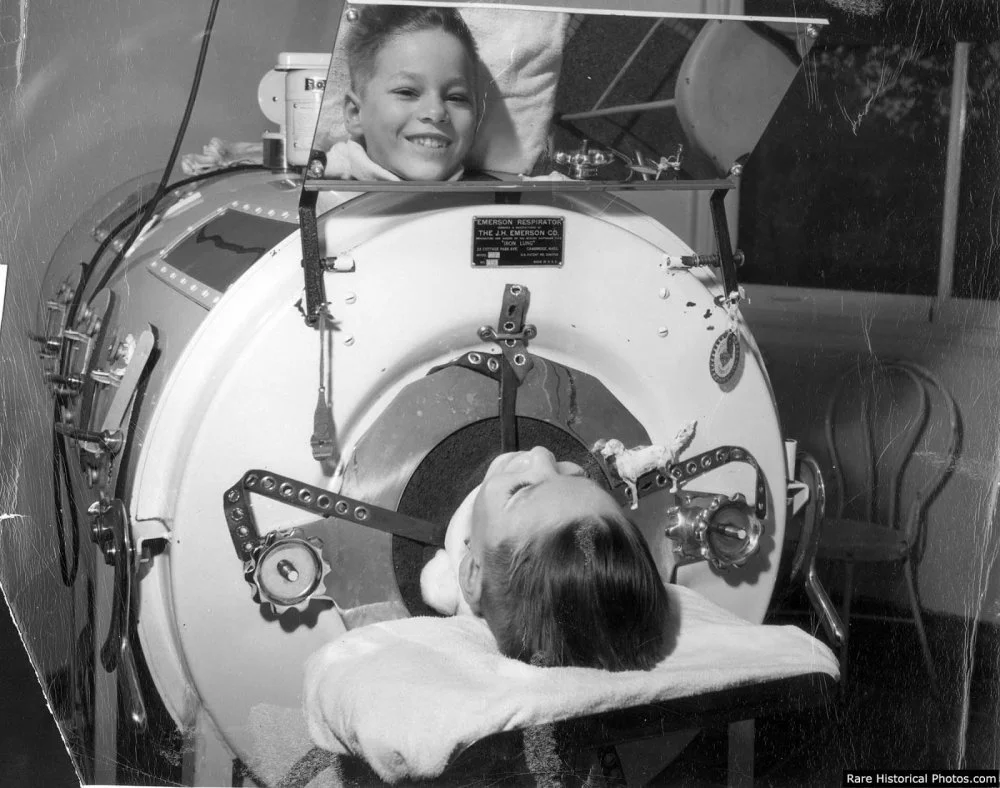 An archival image of a young boy smiling at the camera through a mirror attached to the top of his iron lung