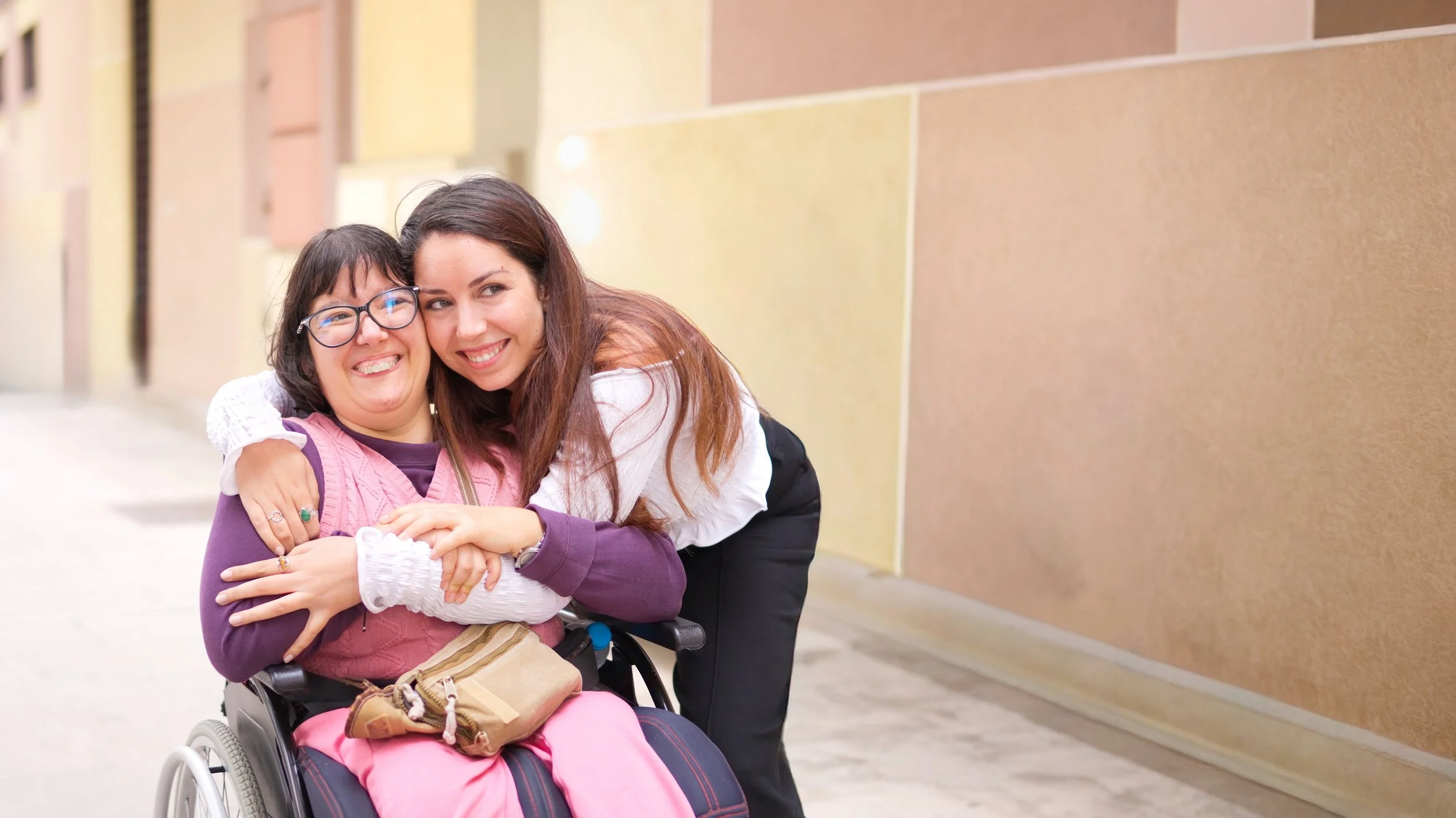 Two smiling women embrace in front of a tan building wall. One woman with dark hair and glasses is sitting in a wheelchair. The other woman stands behind her with her arms around her.