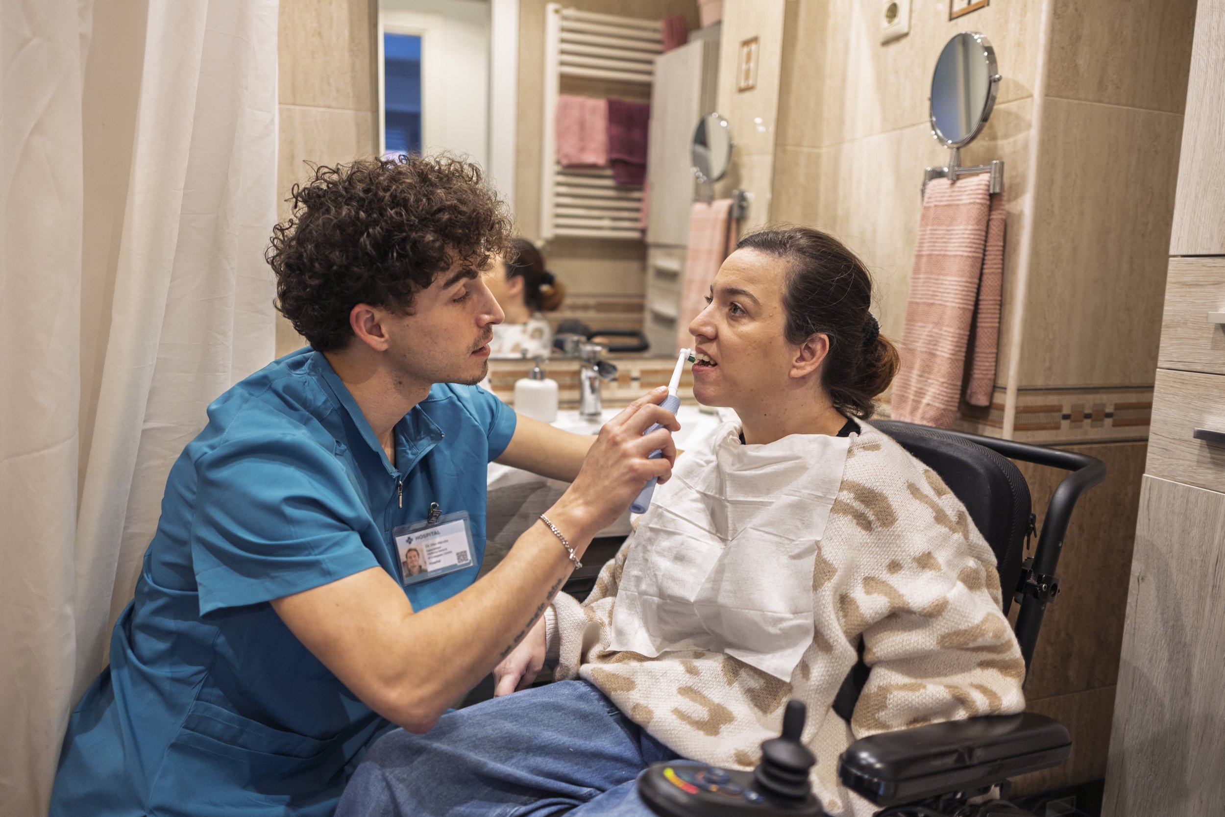 In a brightly lit bathroom, a dental assistant with curly hair, wearing blue scrubs, is assisting a woman with an electric toothbrush. The woman, seated in a wheelchair, is wearing a patterned sweater.