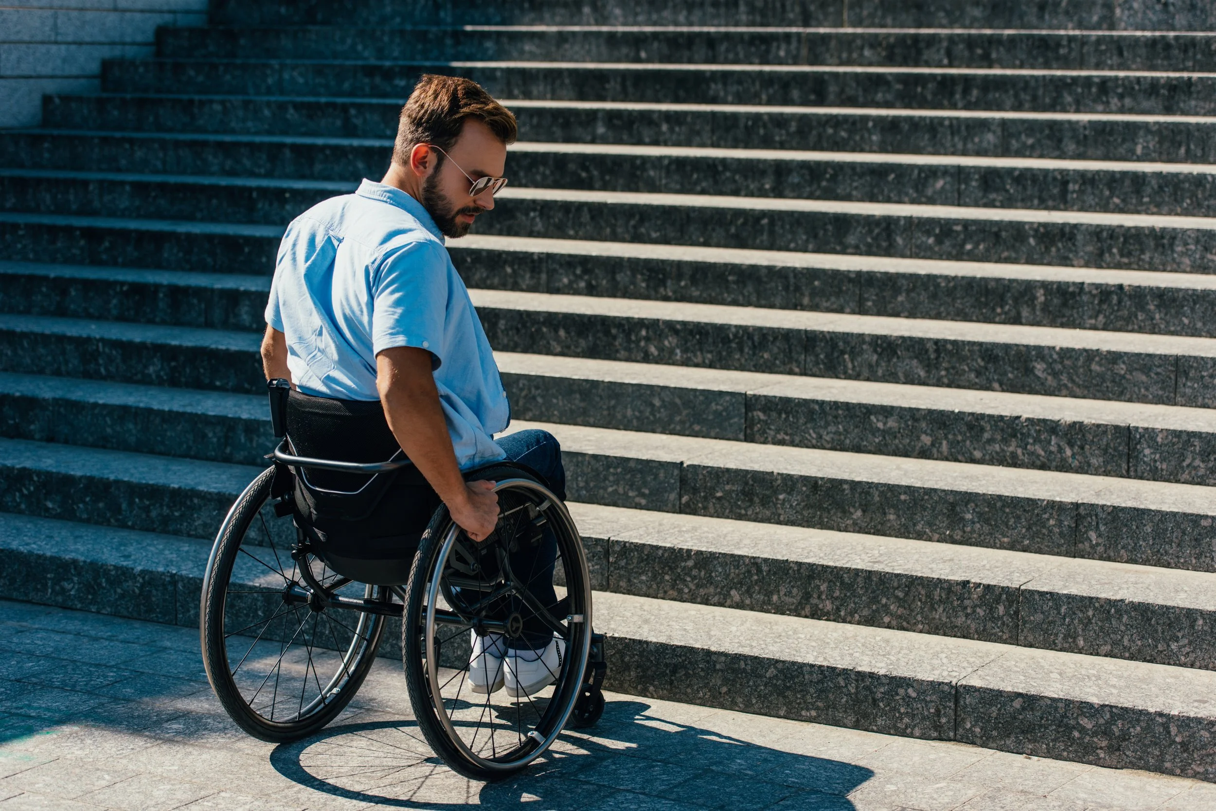 A man with short brown hair and a beard is seated in a black wheelchair outside on a sunny day, facing away from the camera and toward a large set of concrete steps. He is wearing a short-sleeved light blue button-down shirt, blue jeans, and glasses.