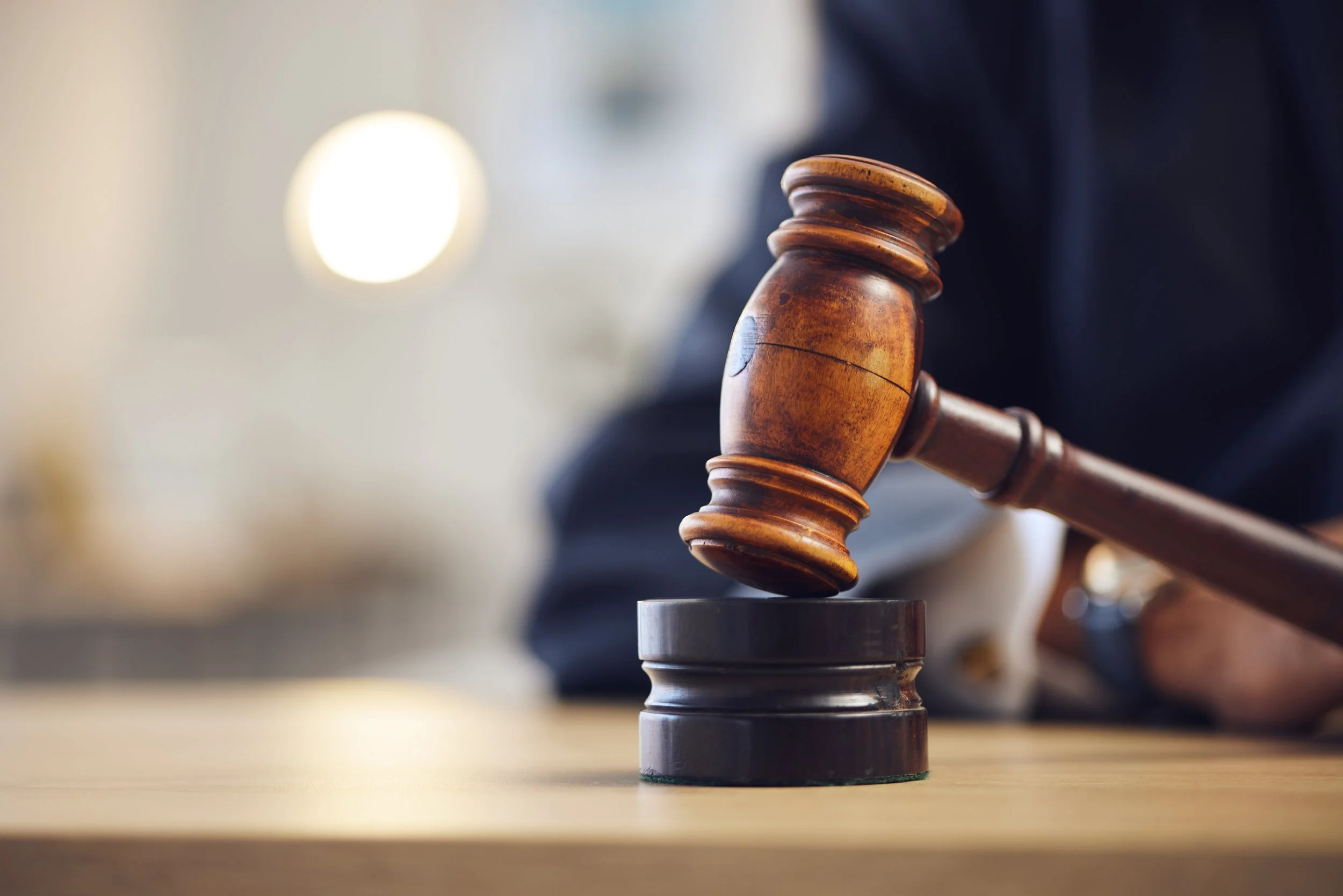 A wooden gavel rests on a dark sound block atop a light wooden table. The gavel handle extends to the right, over a blurred adult dressed in a dark suit and white shirt cuffs, who is seated in the background.