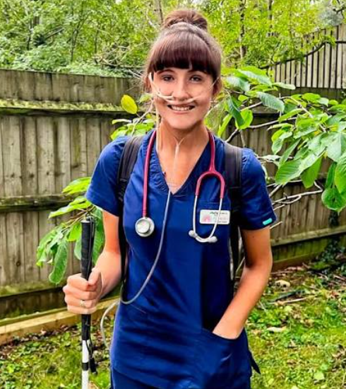 Alexandria in her scrubs and stethoscope, connected to an oxygen tank, holding her cane.