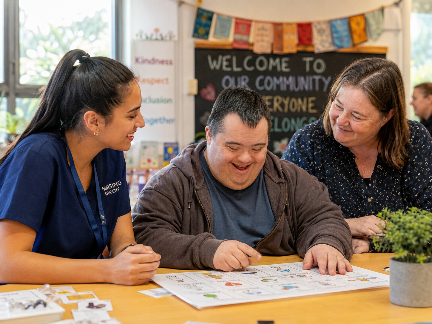 A nursing student and a woman support an adult with Down syndrome as he uses a visual activity guide at a table in a welcoming community setting.