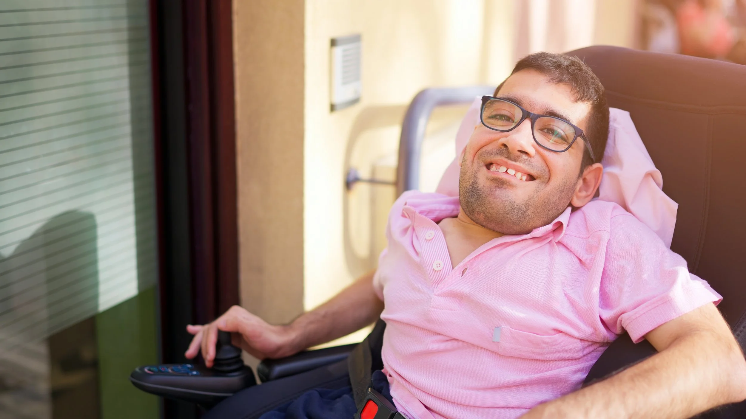 A smiling man with dark hair and glasses is sitting in a motorized wheelchair indoors. He is wearing a pink polo shirt and a seatbelt.
