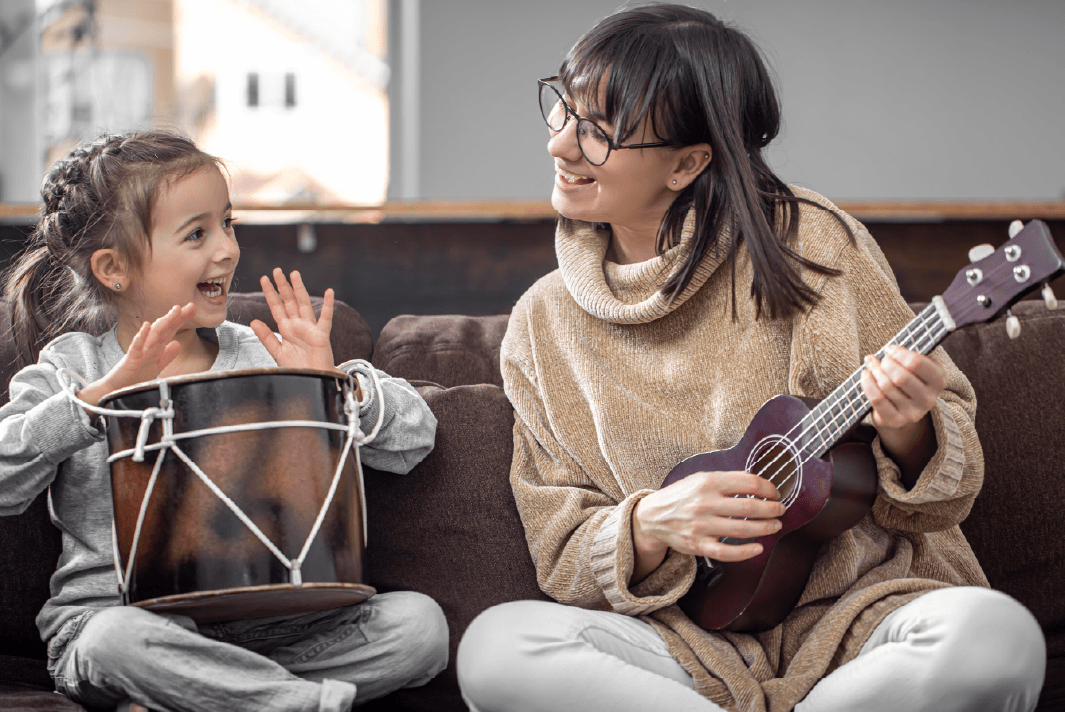 A young child sits cross-legged on a brown sofa, smiling and playing a small drum, with her hands raised in excitement. Beside her, a young adult woman with dark hair and glasses is playing a ukulele, smiling as she watches the child.