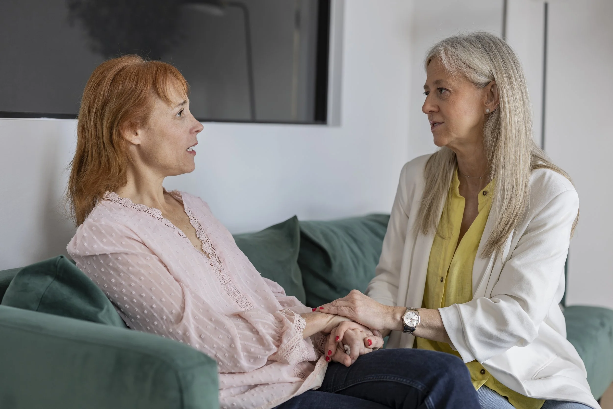 Two women sit close together on a green couch while talking.