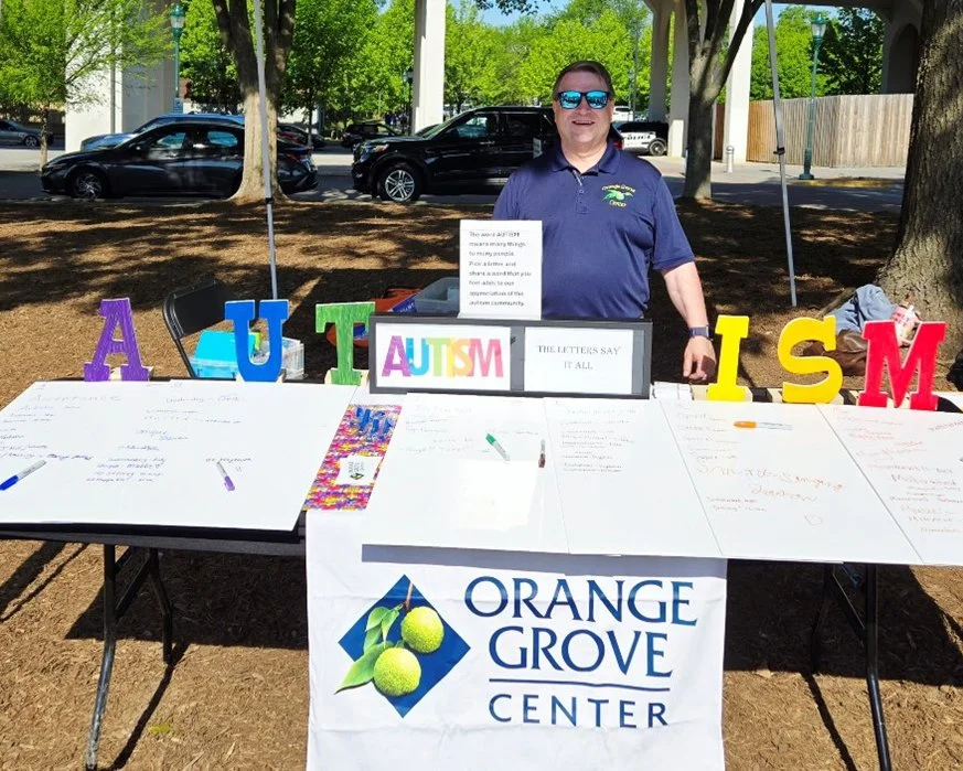 David Cox stands behind an Orange Grove Center table at the Chattanooga Autism Walk, where colorful letters spelling “AUTISM” and poster boards invite participants to write words they associate with autism.