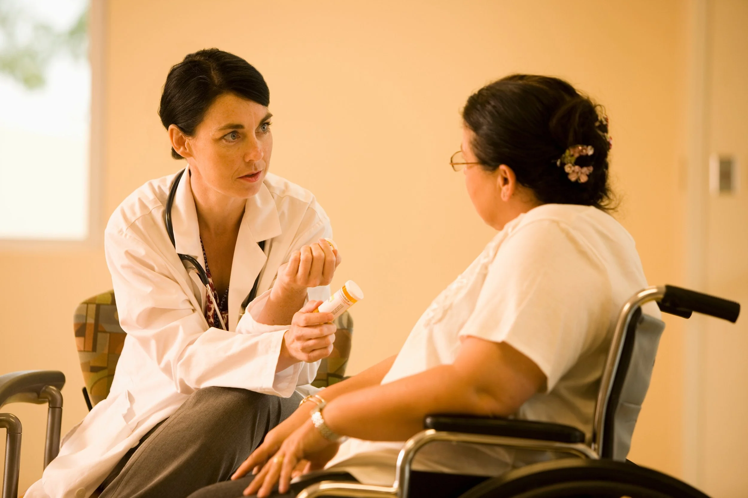 A woman in a white coat and stethoscope sits and talks with a patient in a wheelchair. The woman in the white coat holds a small bottle. The patient has her hair up with an ornamental clip. The setting is indoors, with natural light from a window.