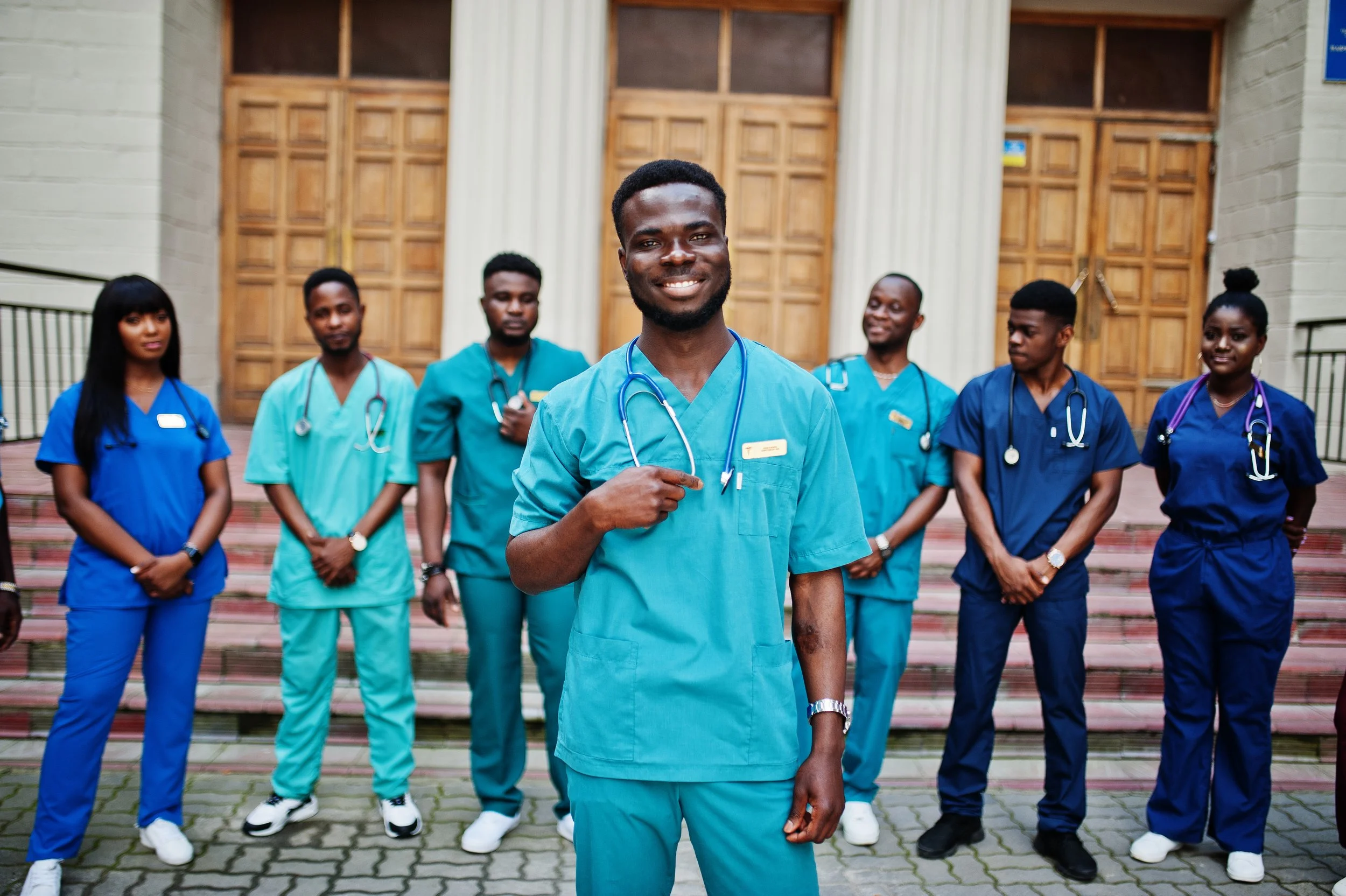 Seven smiling Africans in blue and green scrubs stand together in front of a brick building.