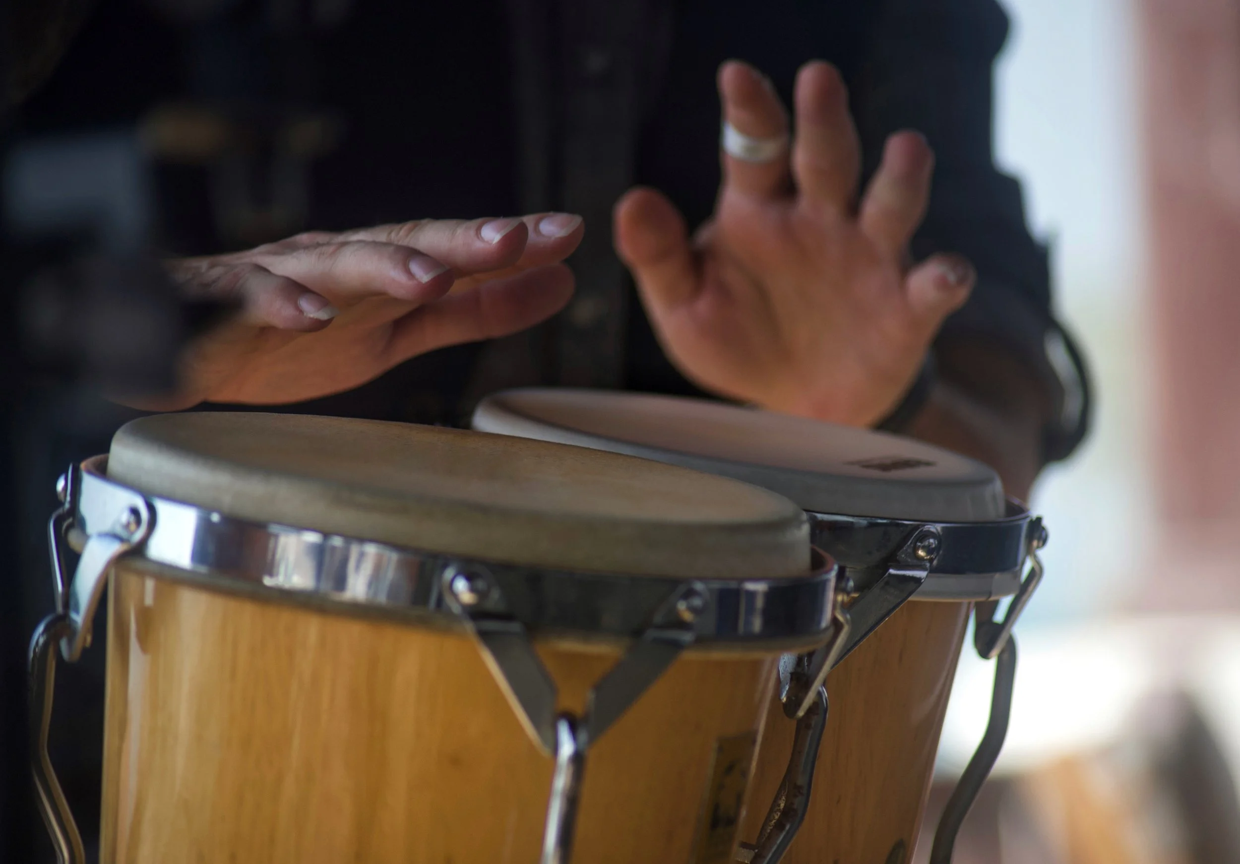 This close up photograph features the hands of a person playing a set of wooden bongos. The hands are poised over the drums, ready to strike.