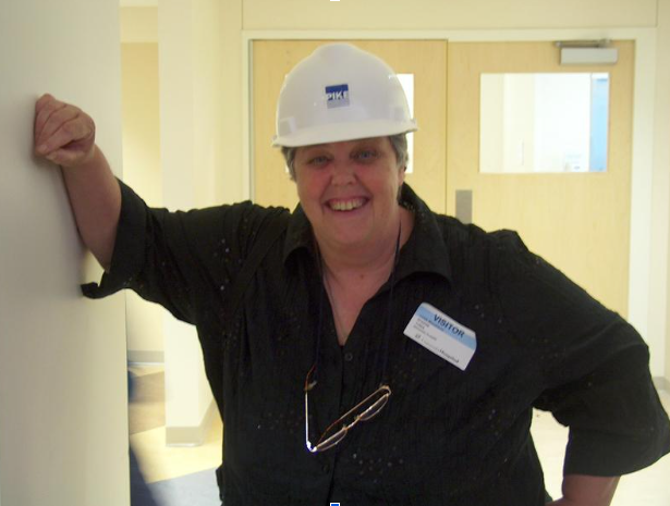 A photo of Linda wearing a hard hat and leaning against a wall with a bright smile on her face.