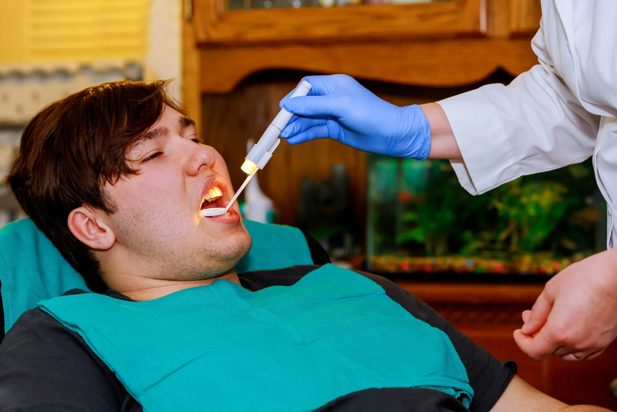 A young adult man lies back in a dental chair with his mouth open wide, illuminated by a small light attached to a dental mirror. A gloved hand holds the dental instrument. He is wearing a dark shirt with a turquoise patient covering.