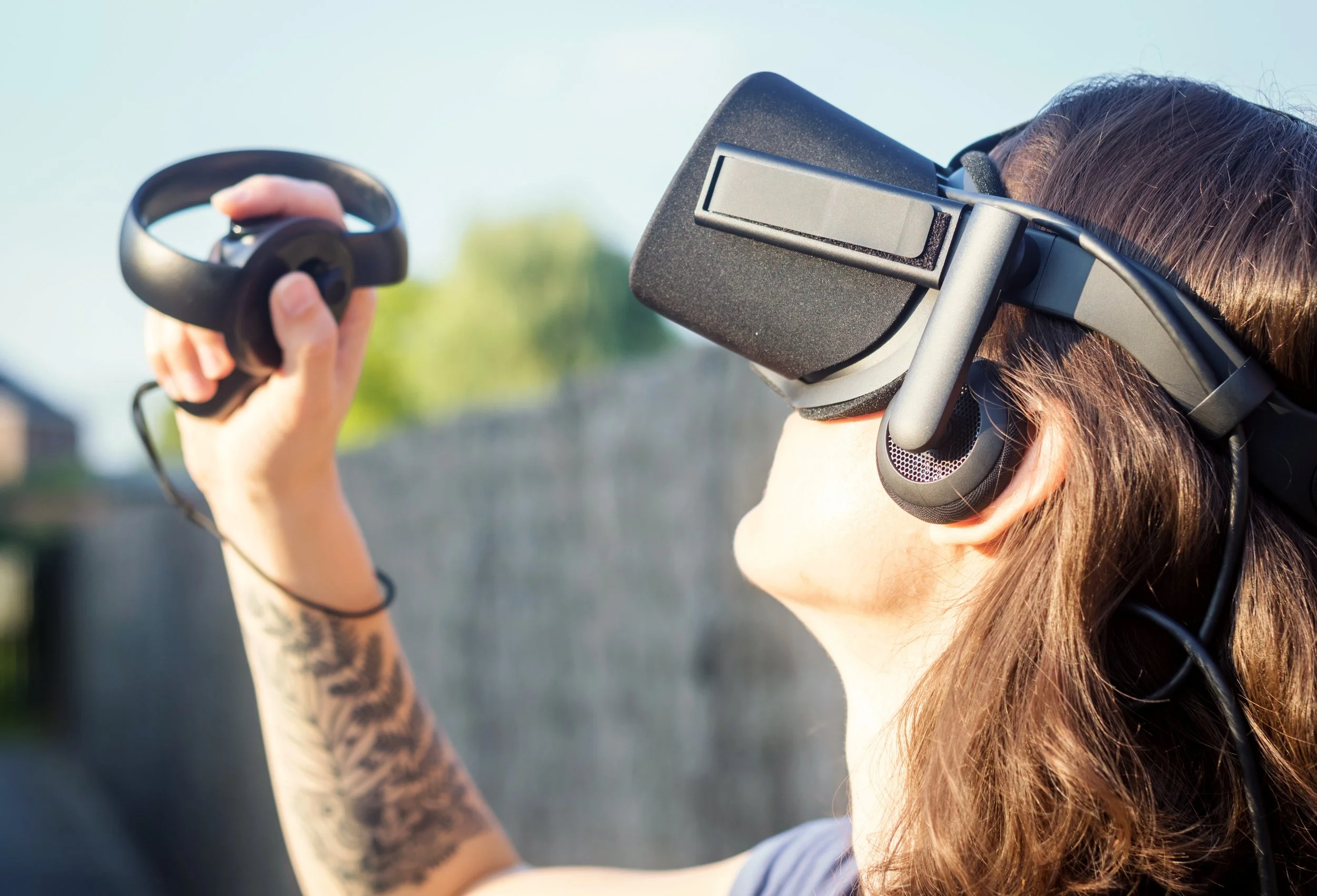 A woman with wavy brown hair is using a virtual reality headset in an outdoor setting during daytime. The woman is seen waist up, looking up towards the blue sky. Her arm is visible and shows a tattoo.