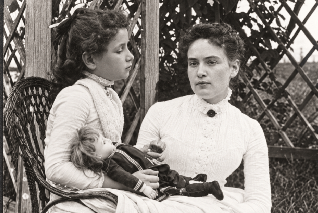 Helen Keller sits outside with her doll in her lap and Annie Sullivan sitting beside her