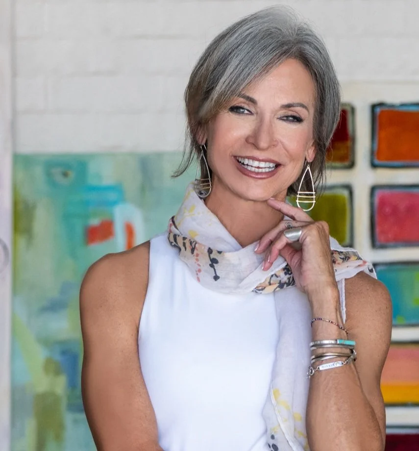 A woman with short gray hair, smiling, wearing a white sleeveless top, patterned scarf, and long earrings, standing in front of a colorful art background.