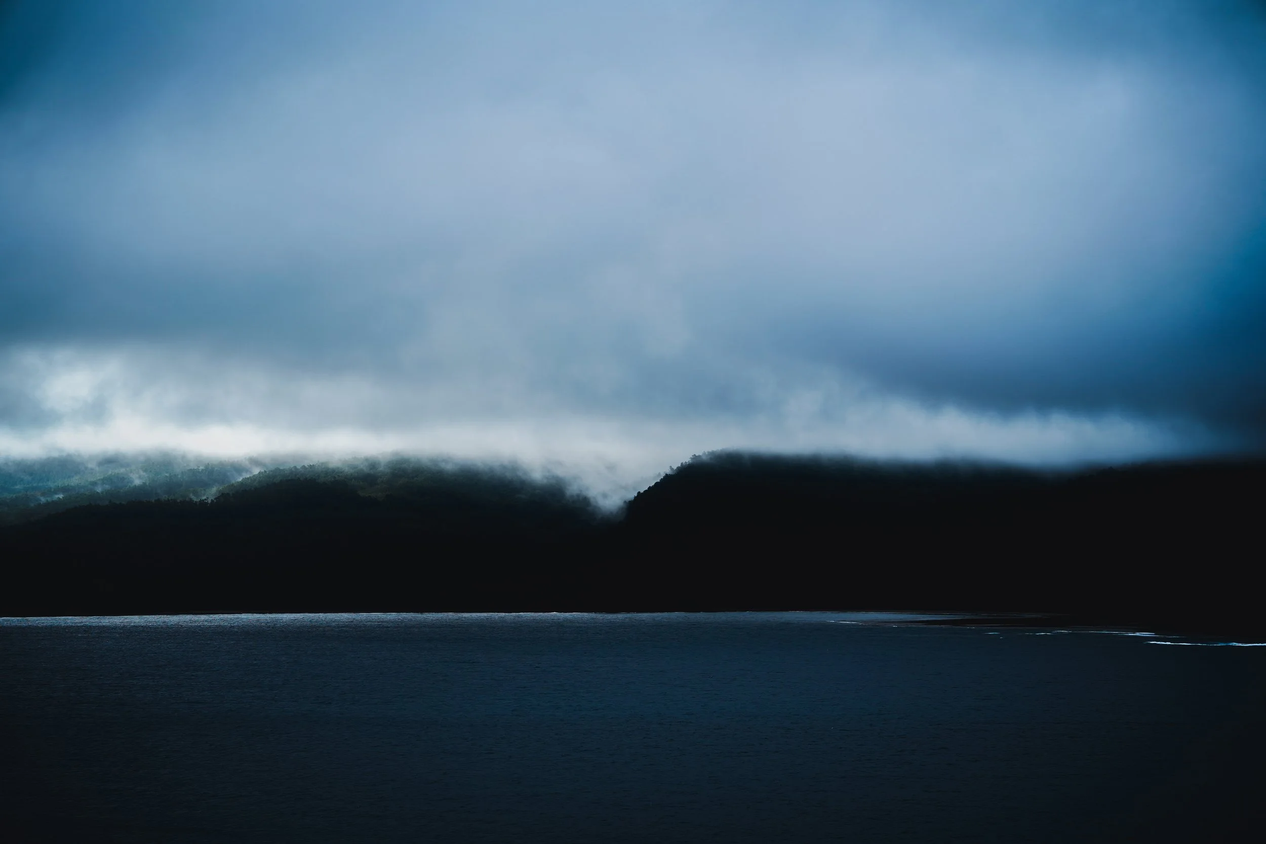 A dark, overcast sky with heavy clouds above a mountain range and a body of water in the foreground.