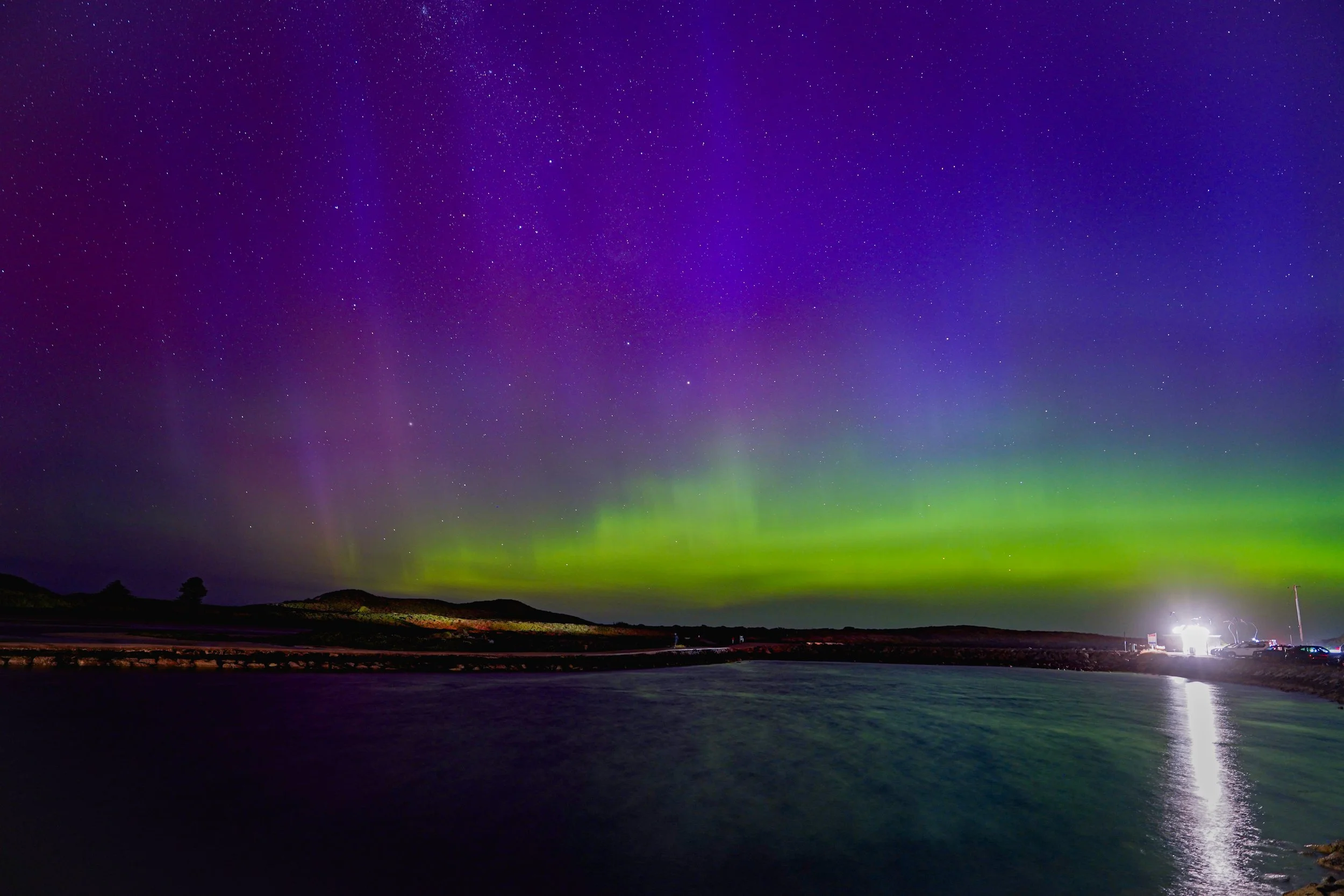 Night sky illuminated by colorful northern lights over a body of water with a few silhouetted trees and distant lights on the shoreline.