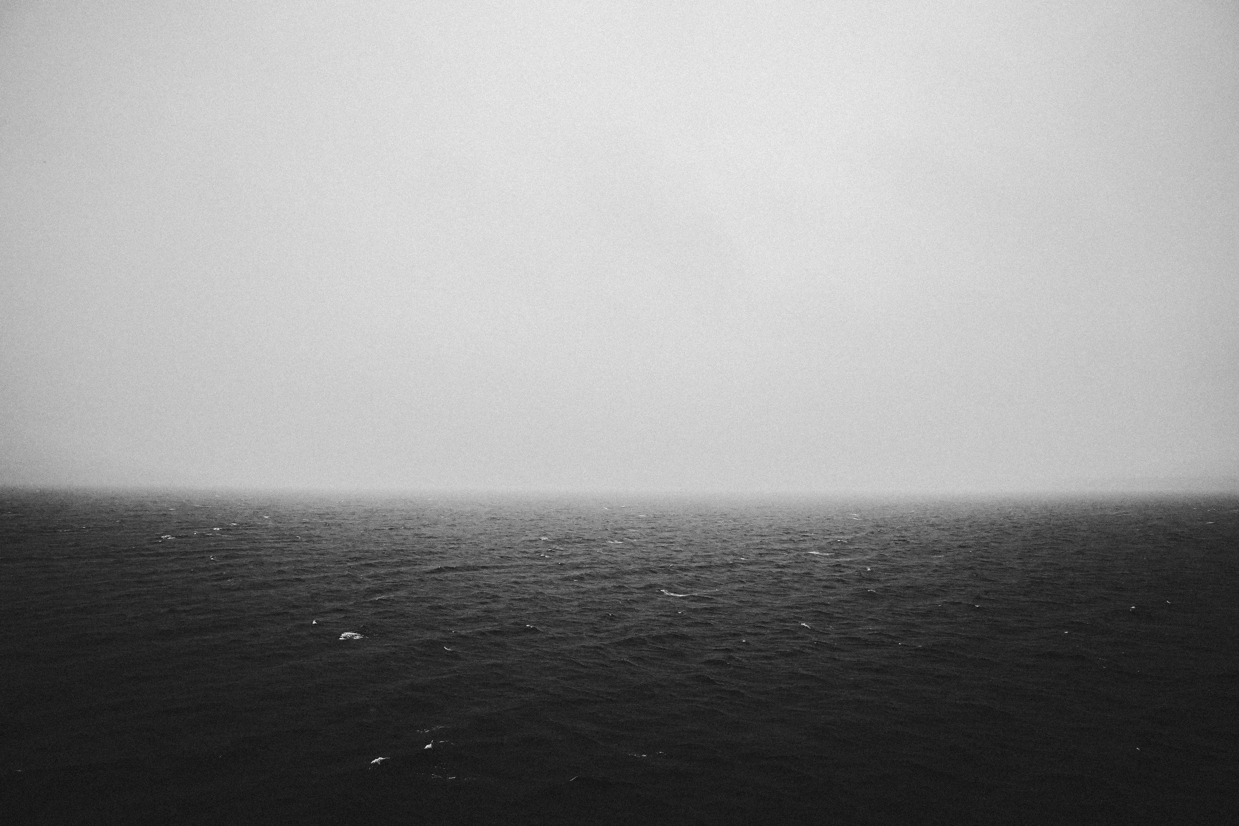 A black and white photograph of the ocean with gentle waves and a distant horizon under a calm sky.