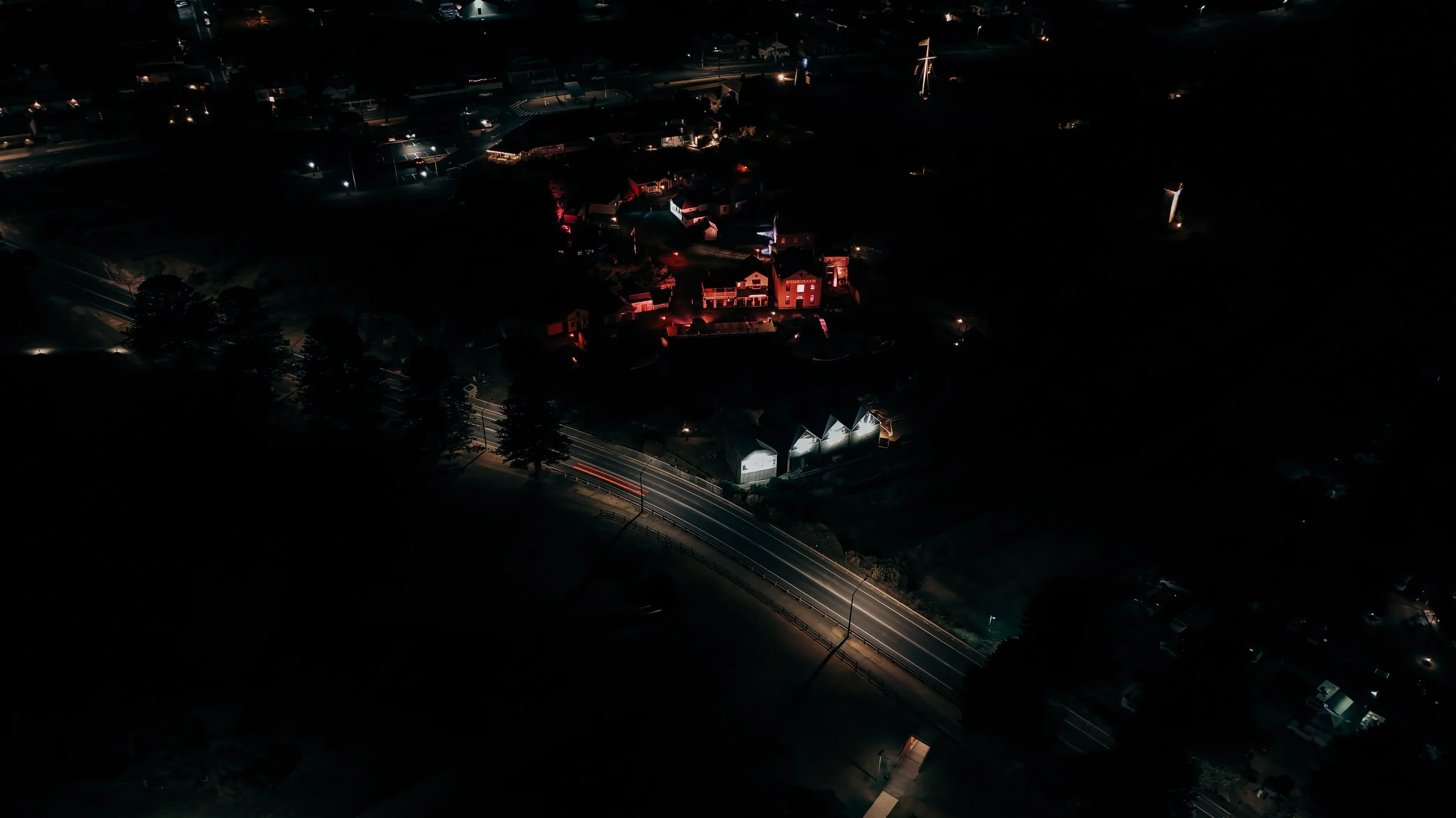 Aerial nighttime view of a neighborhood featuring houses, some with external lighting, and a winding road with car light streaks.