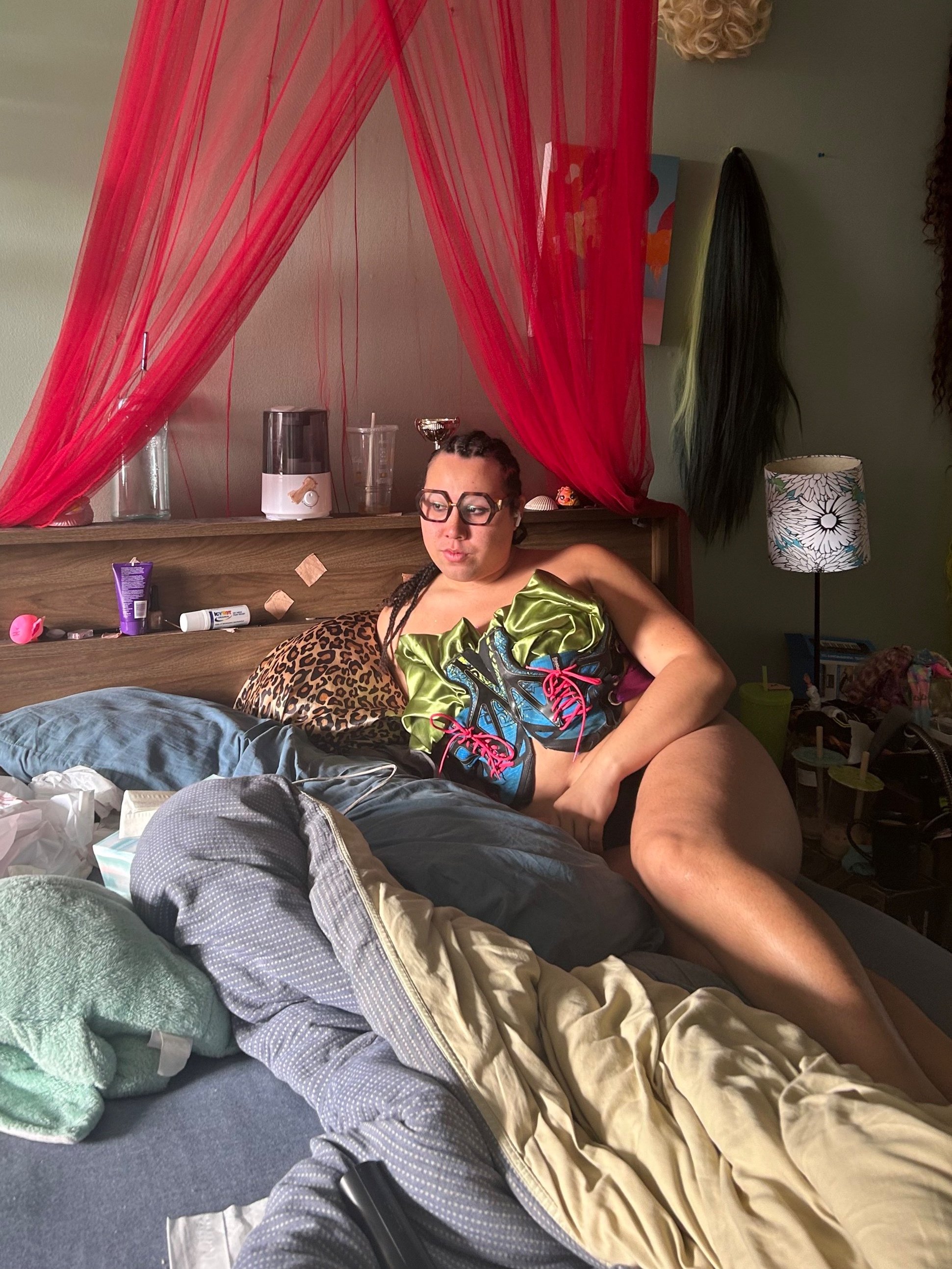 Picture of a model laying in bed.Deconstructed blue sneakers reveal satin green ruffling fabric cover their chest. They are gazing towards the sun, wearing glasses and braided hair.
