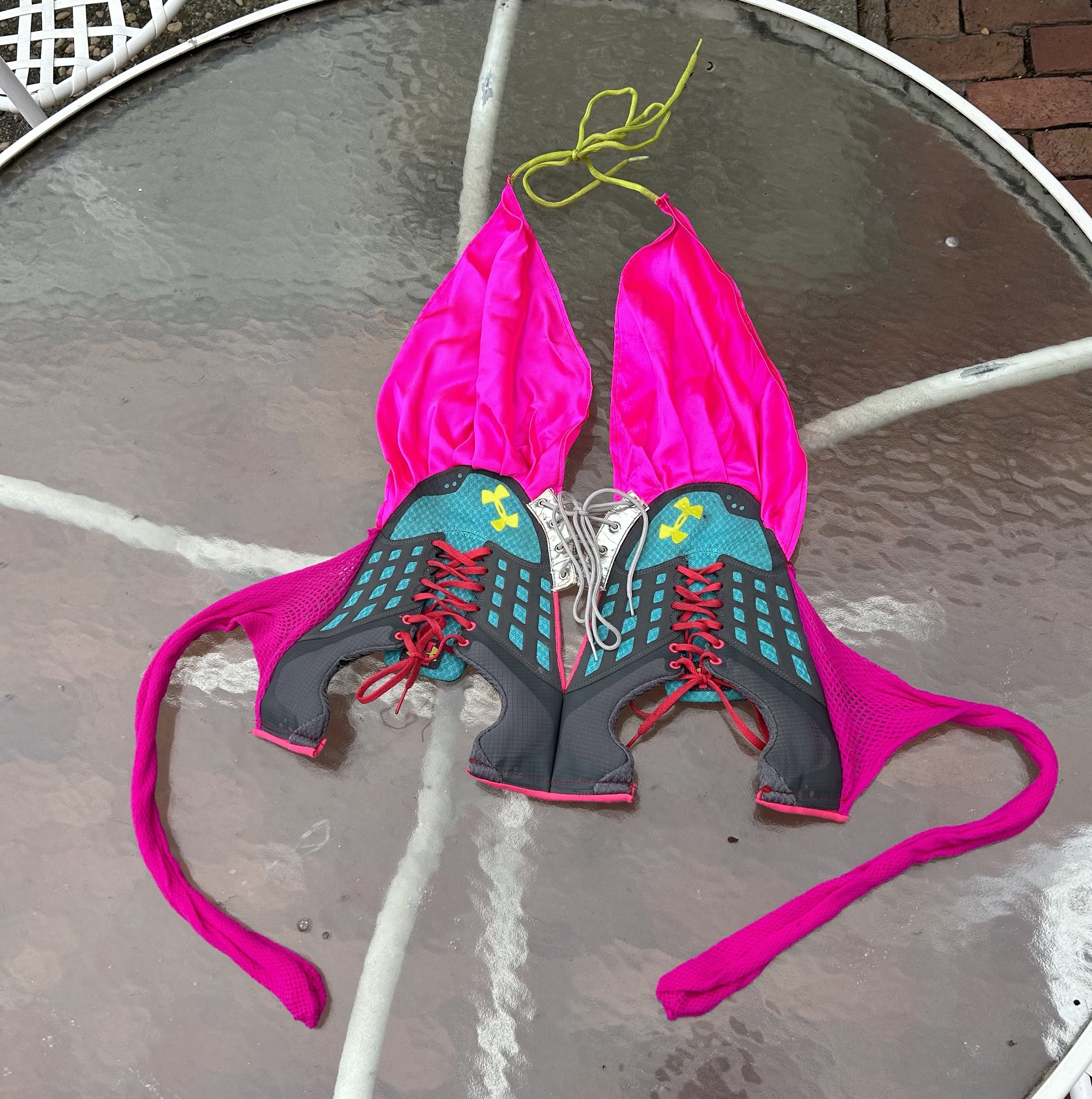 Hot pink silk straps adorn teal deconstructed sneakers that form a bra shape. Red shoelaces, a neon green neck strap, and metallic silver laces are all part of the sneaker top.