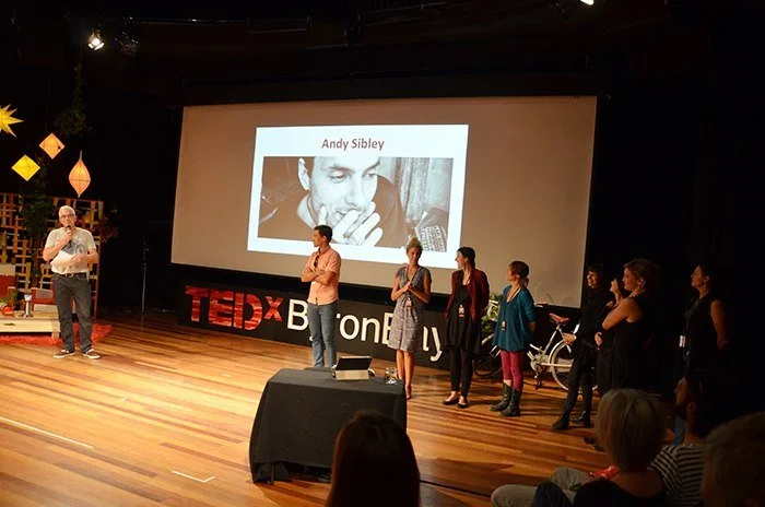 A group of people on stage at a TEDx event, with a large screen displaying a black and white photo of Andy Sibley. The stage has TEDx branding, and the audience is seated in front.