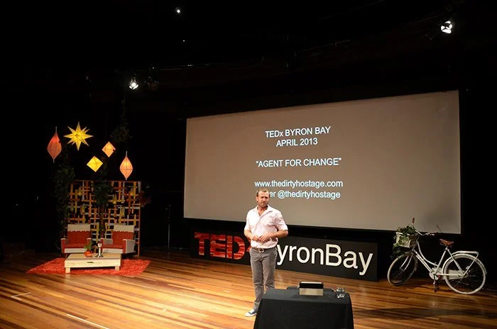 A man is standing on a stage with a TEDx Byron Bay banner, giving a presentation titled 'Agent for Change' in April 2013. The backdrop includes a large screen displaying event details, decorative hanging lamps, a bicycle, and a seating area with chairs and a small table.