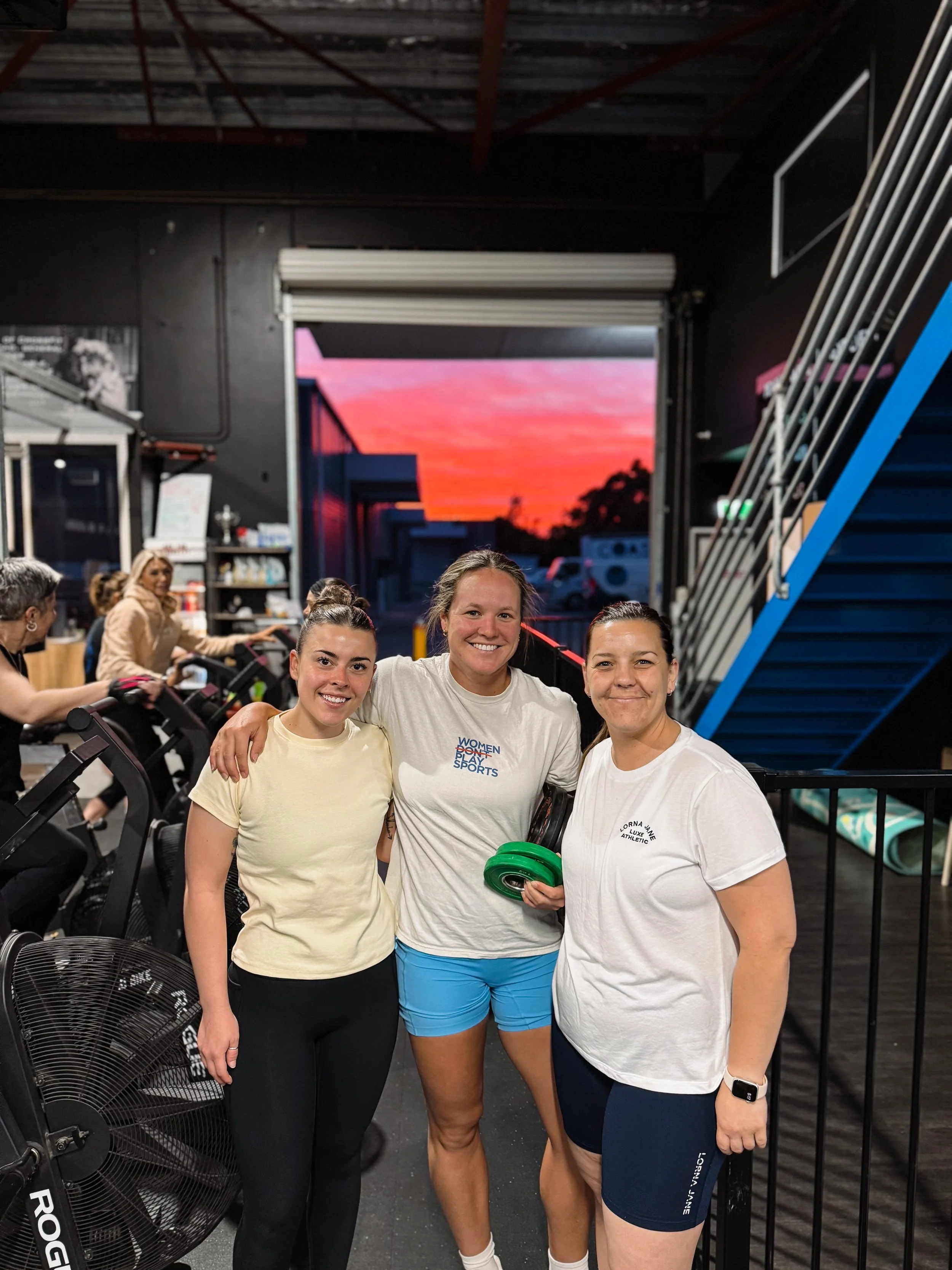 Three women smiling inside a gym (Evolve Fitness Collective), standing together with their arms around each other. In the background, other members of the community are working out, and there is a large window with a sunset sky visible.