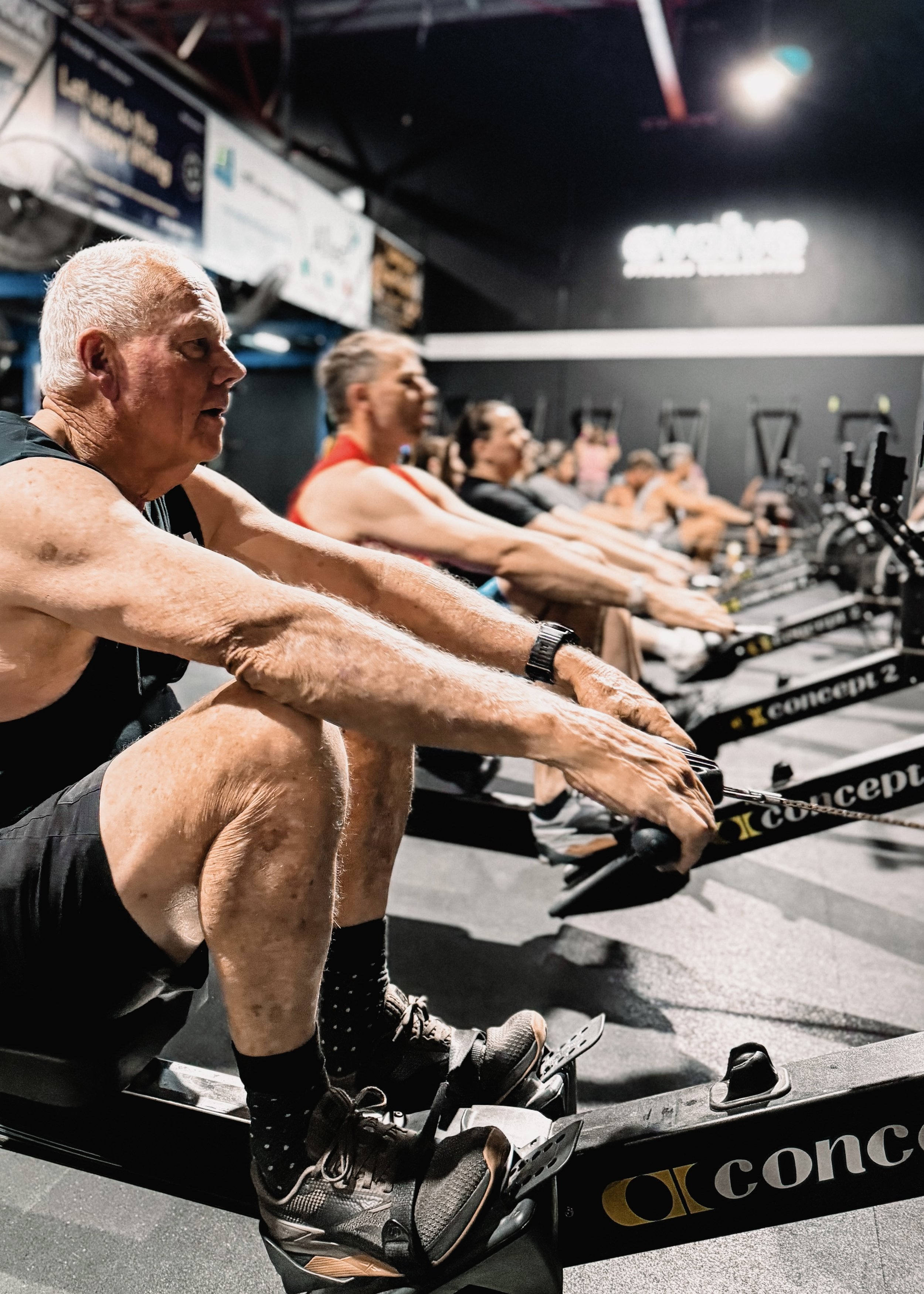Older man exercising on a rowing machine in a gym with other people in the background.