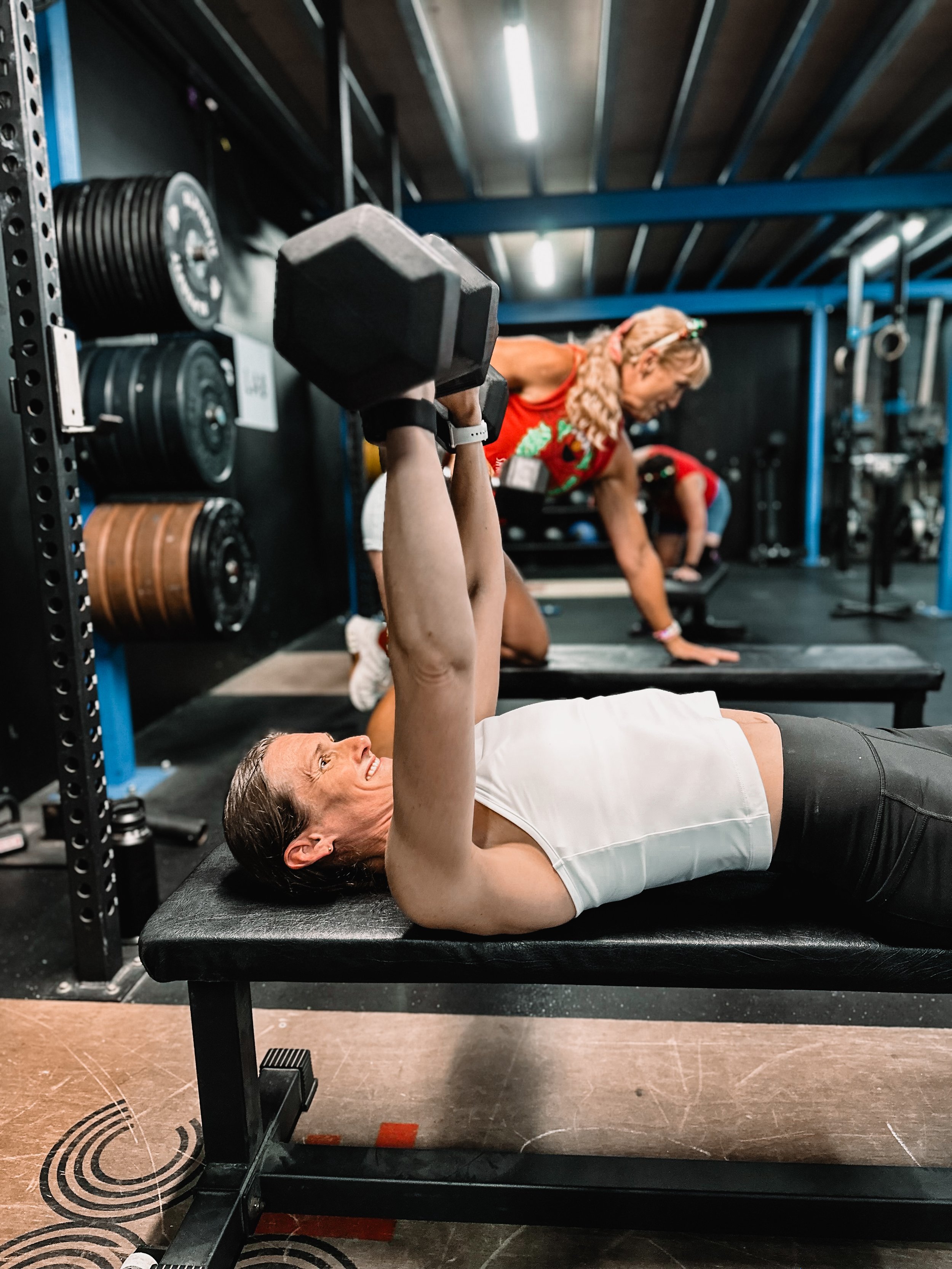 A woman is lying on a workout bench and lifting a dumbbell in a gym with lots of exercising going on around.