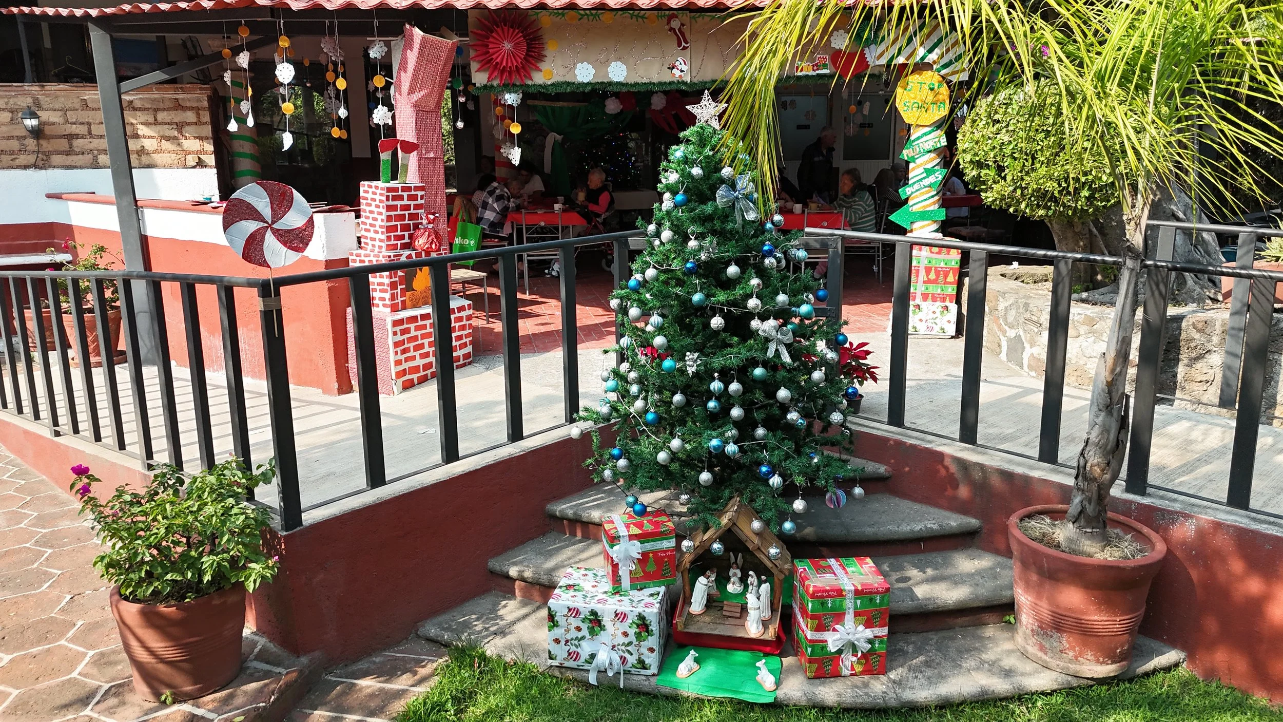 Arbol de Navidad decorado con esferas plateadas y azules, rodeado de regalos envueltos en papel navideño. En el fondo, espacio decorado con adornos navideños y mesas con personas sentadas, en un ambiente festivo al aire libre.