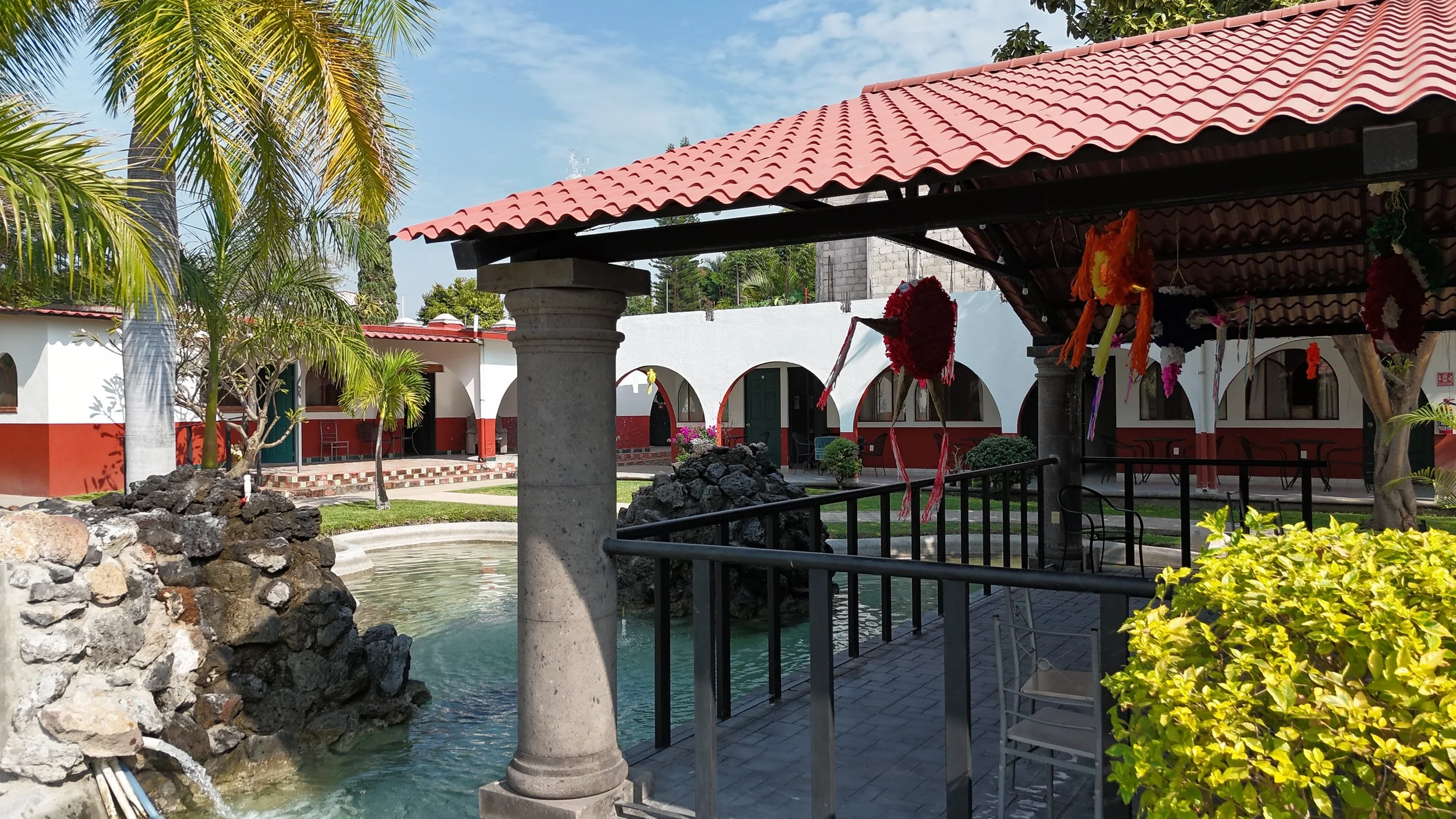 Un patio con fuente de agua, columnas de concreto y techo de tejas rojas, rodeado de plantas y construcciones blancas con arcos, decorado con piñatas de colores.