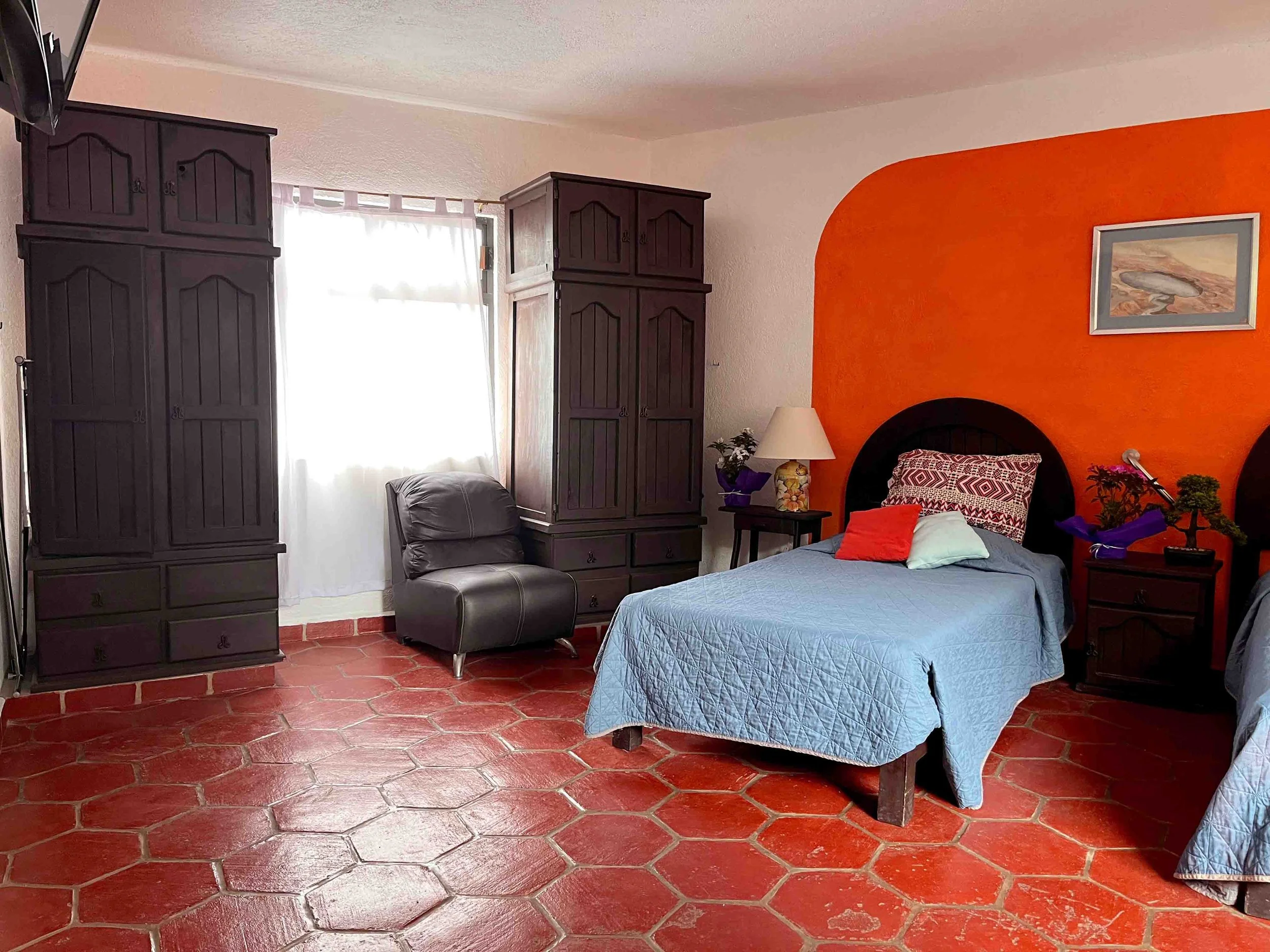 Habitación con una cama doble con cubrecama azul, cabecera negra, una almohada pattern y otra de color sólido, dos mesitas de noche con lámparas y plantas, armarios oscuros y una silla gris, en la pared naranja hay un cuadro.