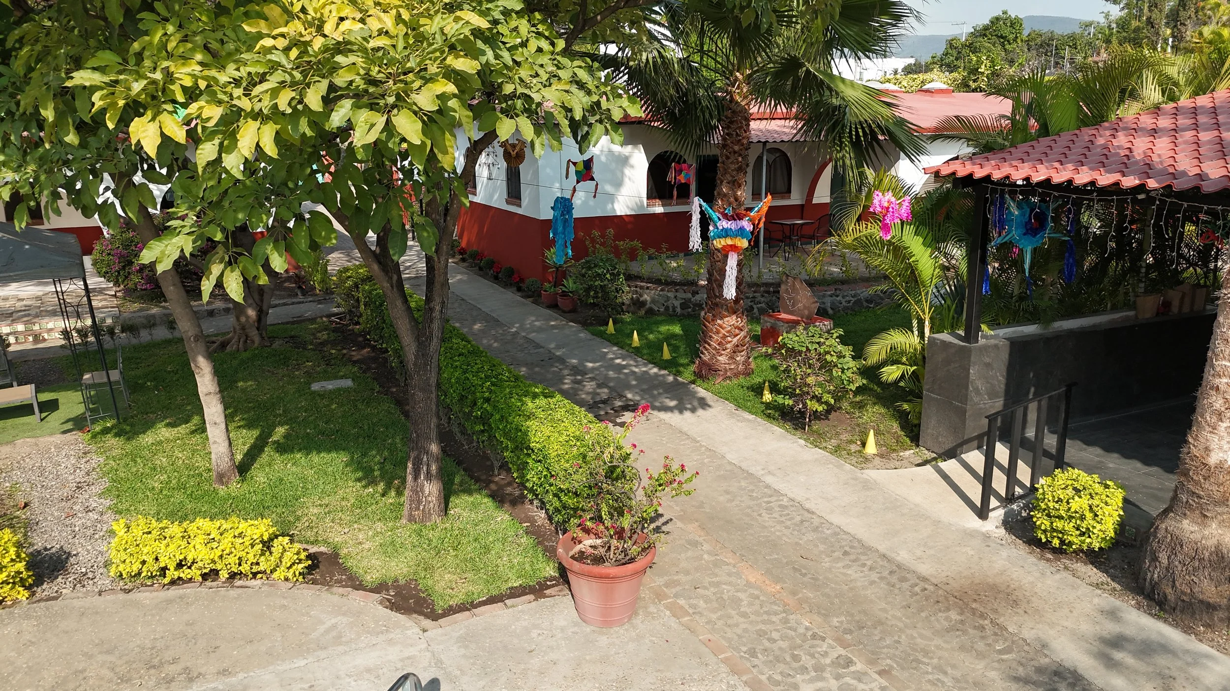 Jardín con árboles, plantas y decoraciones coloridas, estructura con techo de tejas rojas, y un sendero de piedra en un ambiente exterior.