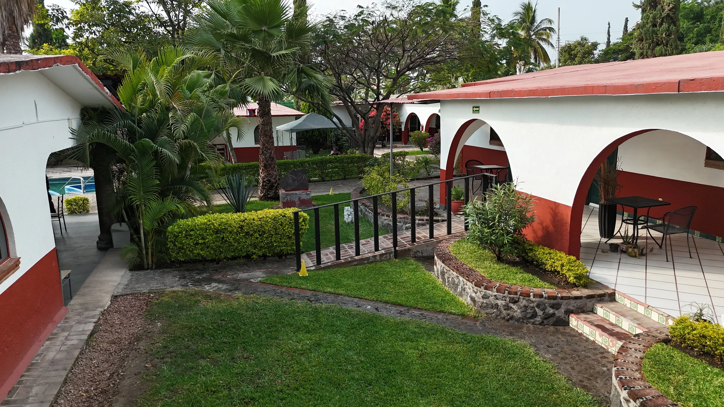 Vista de un área de jardines y habitaciones tipo cabaña con arcos en un complejo turístico, con plantas, caminos y una piscina en el fondo.