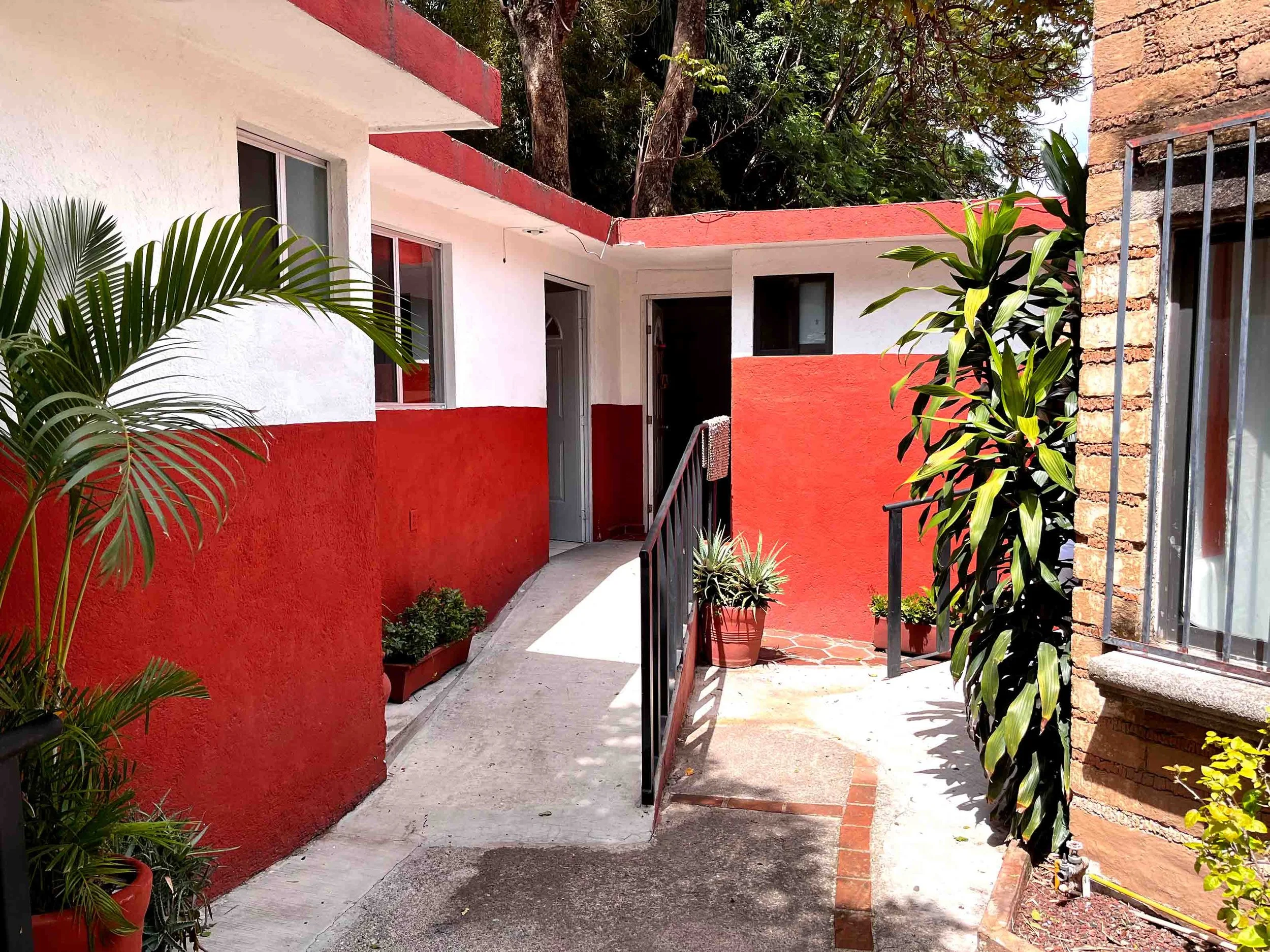 Vista del patio de una casa con paredes pintadas en blanco y rojo, con plantas en macetas y una rampa de acceso.