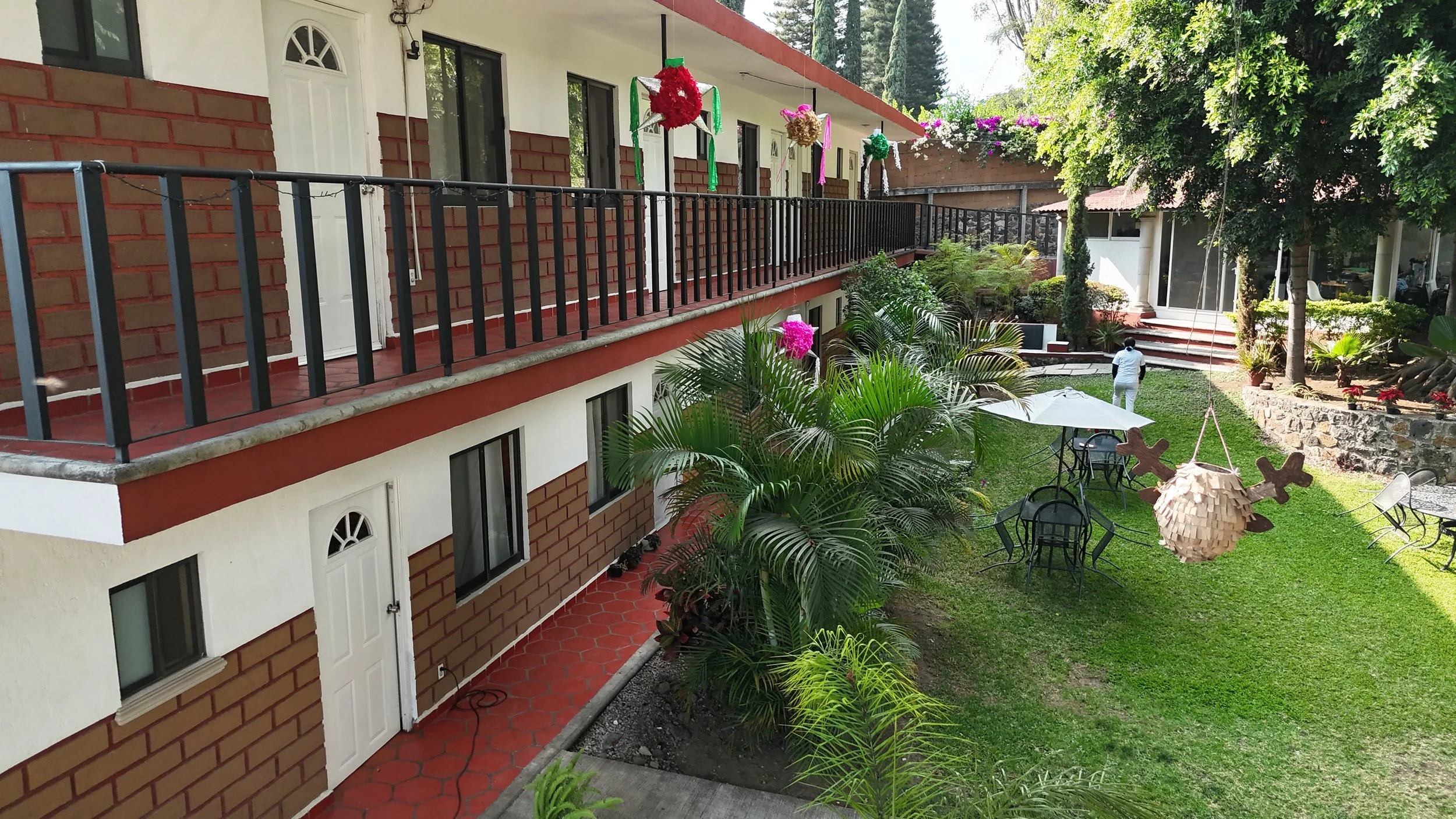 Vista de un jardín con mesas y sillas, palmeras y plantas decorativas, un paraguas blanco y adornos colgantes de colores en una terraza de un edificio de dos pisos, con una mujer caminando y árboles al fondo.