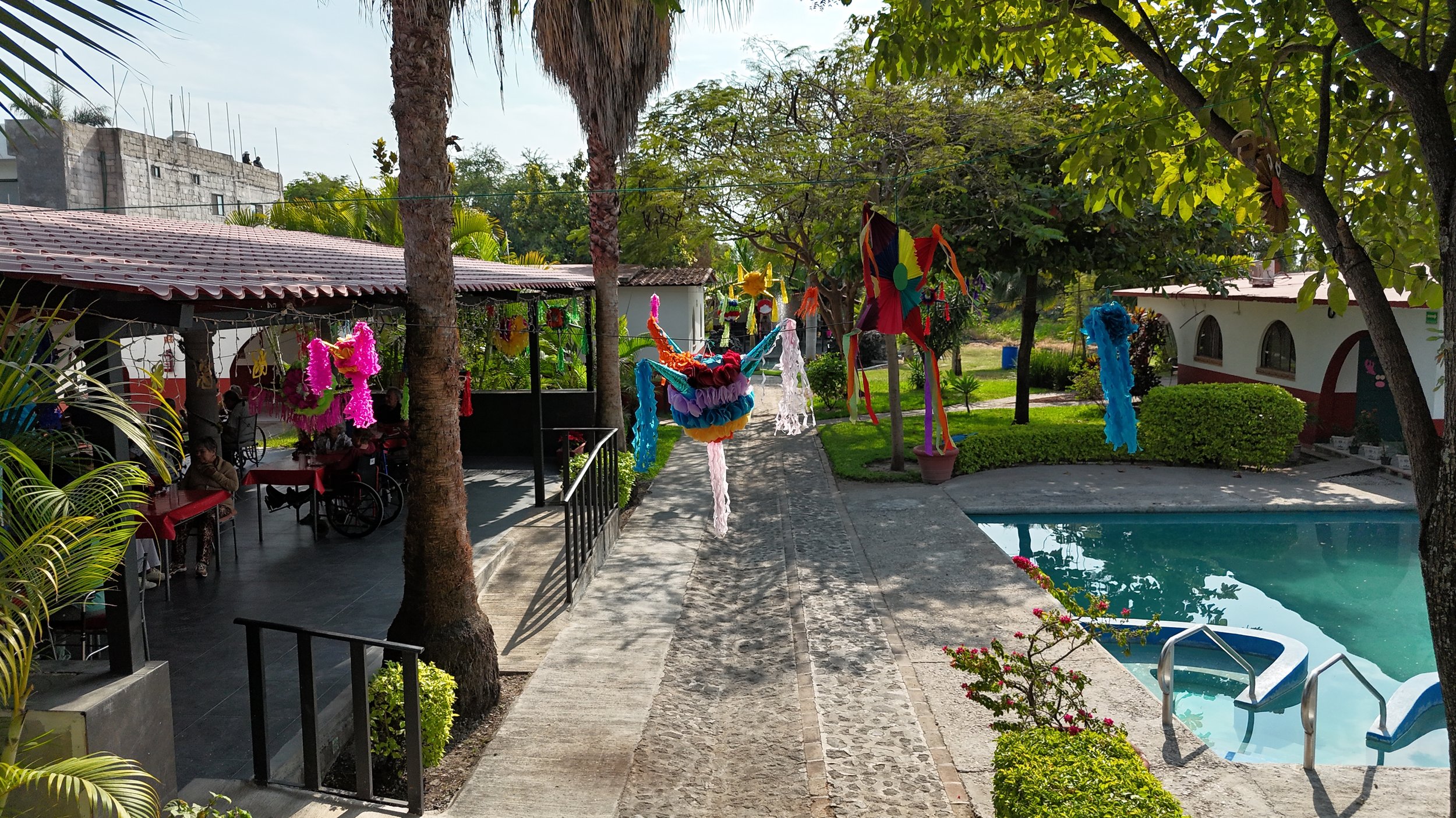 Área de piscina con decoración de papel y tela de colores, árboles y plantas verdes, estructura de techo de tejas a la izquierda, y personas en sillas de ruedas bajo sombra de árboles.