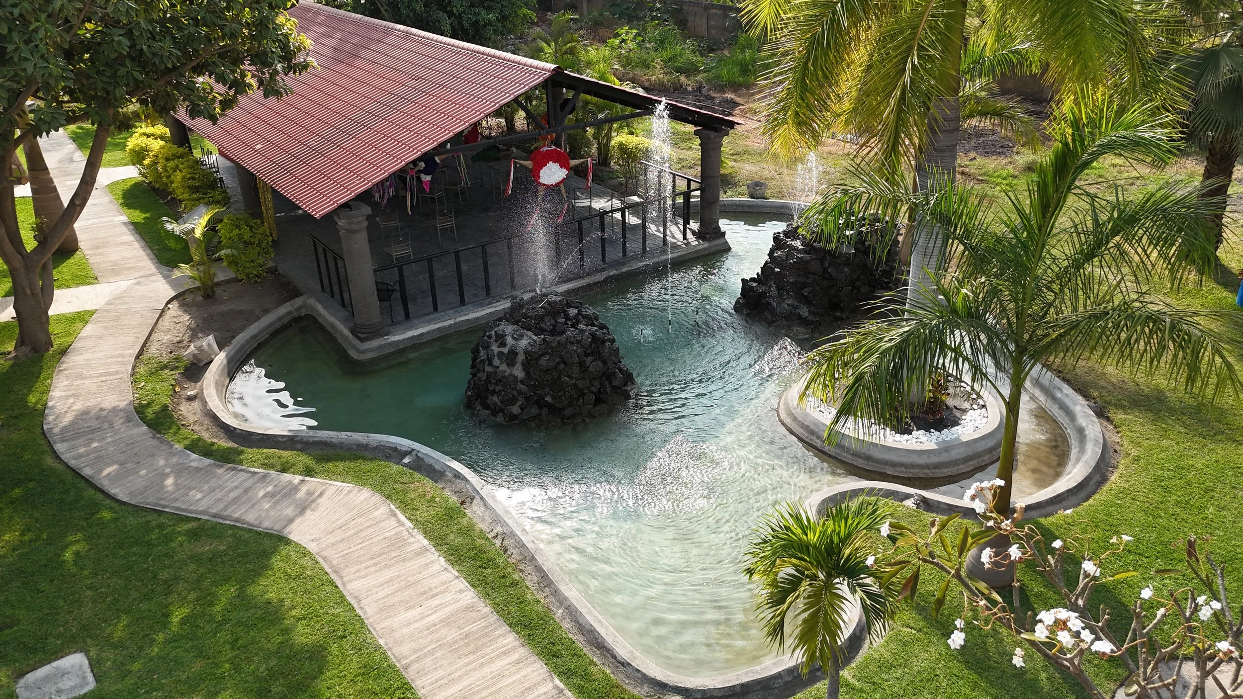 Jardín con fuente de agua, camino pavimentado, árboles y áreas verdes, estructura con techo rojo y columnas delanteras.