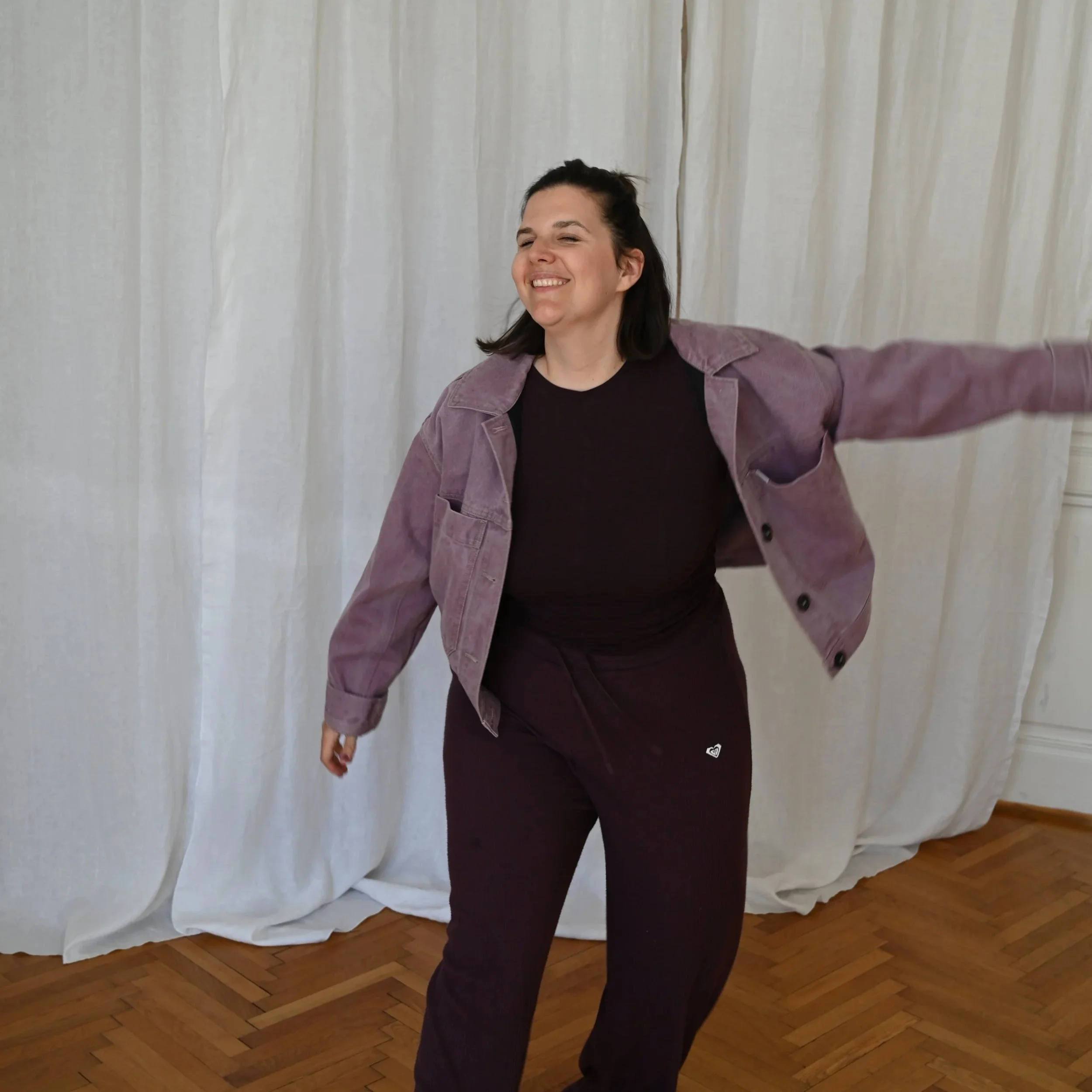Eine lachende Frau in einem lilafarbenen Jacke und schwarzen Shirt, die auf einem Holzparkett steht vor weißen Vorhängen.