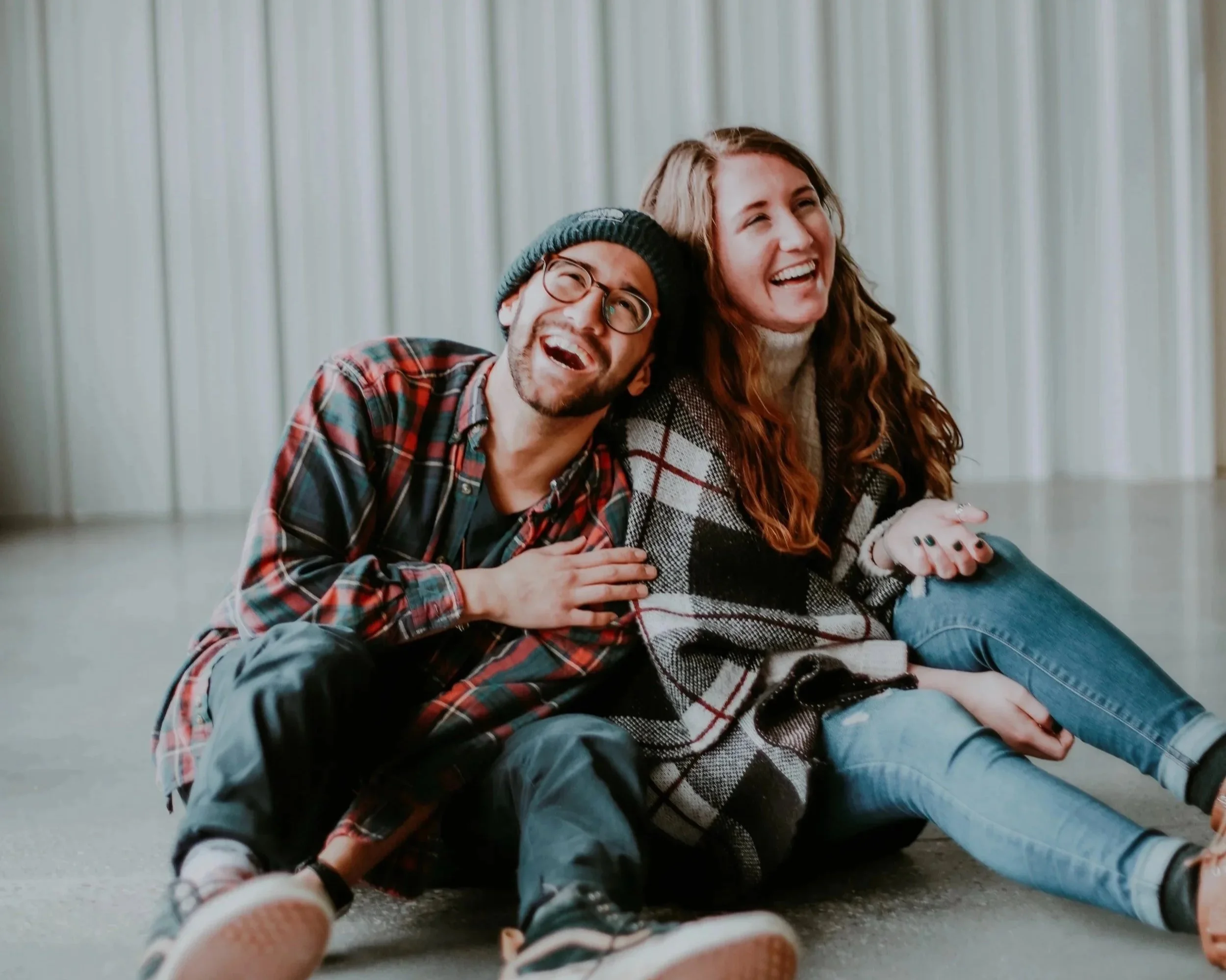 Lachendes, aneinander gelehntes Paar sitzt auf dem Boden, eine männliche und eine weibliche Person, in einem Raum mit weißen Vorhängen im Hintergrund.