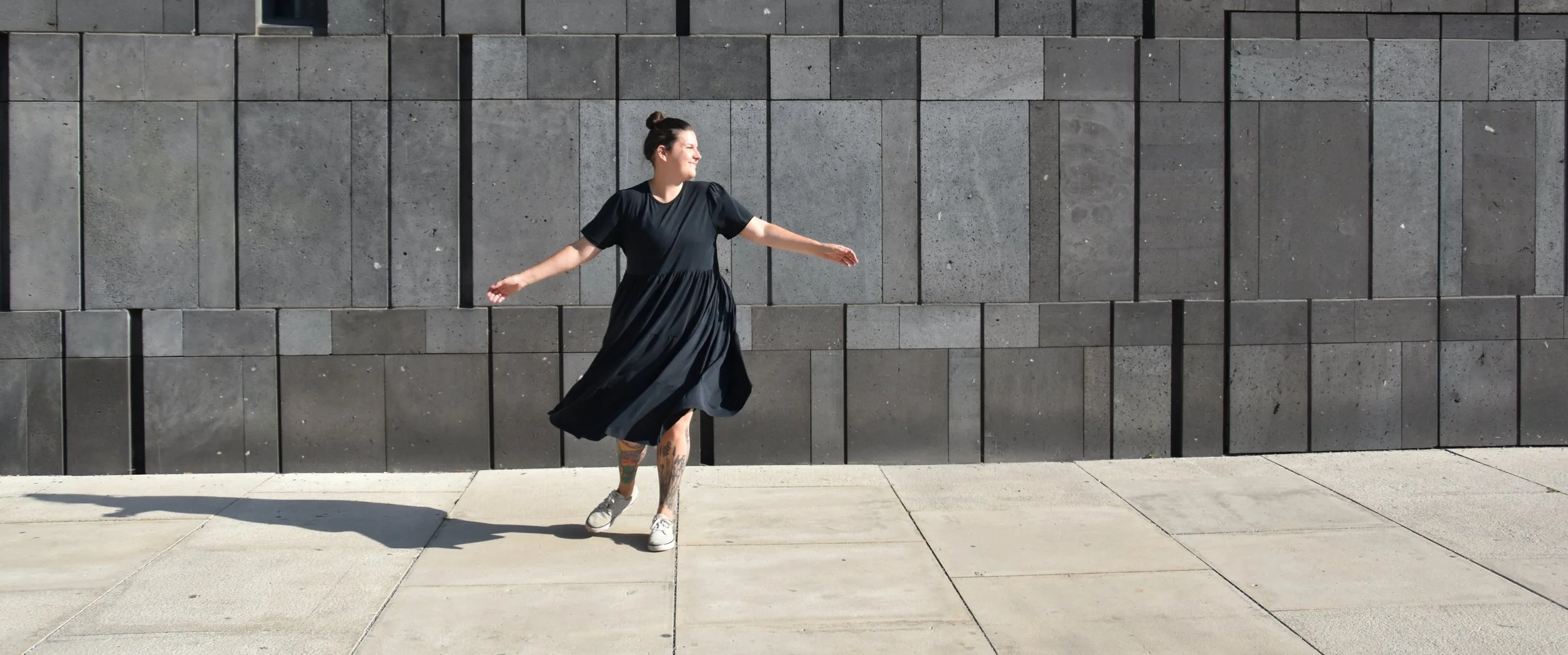 Systemische Coach Isabella Vedernjak tanzt vor einer grauen Steinwand, trägt ein schwarzes Kleid und Sneakers, mit sichtbaren Tattoos auf den Beinen.