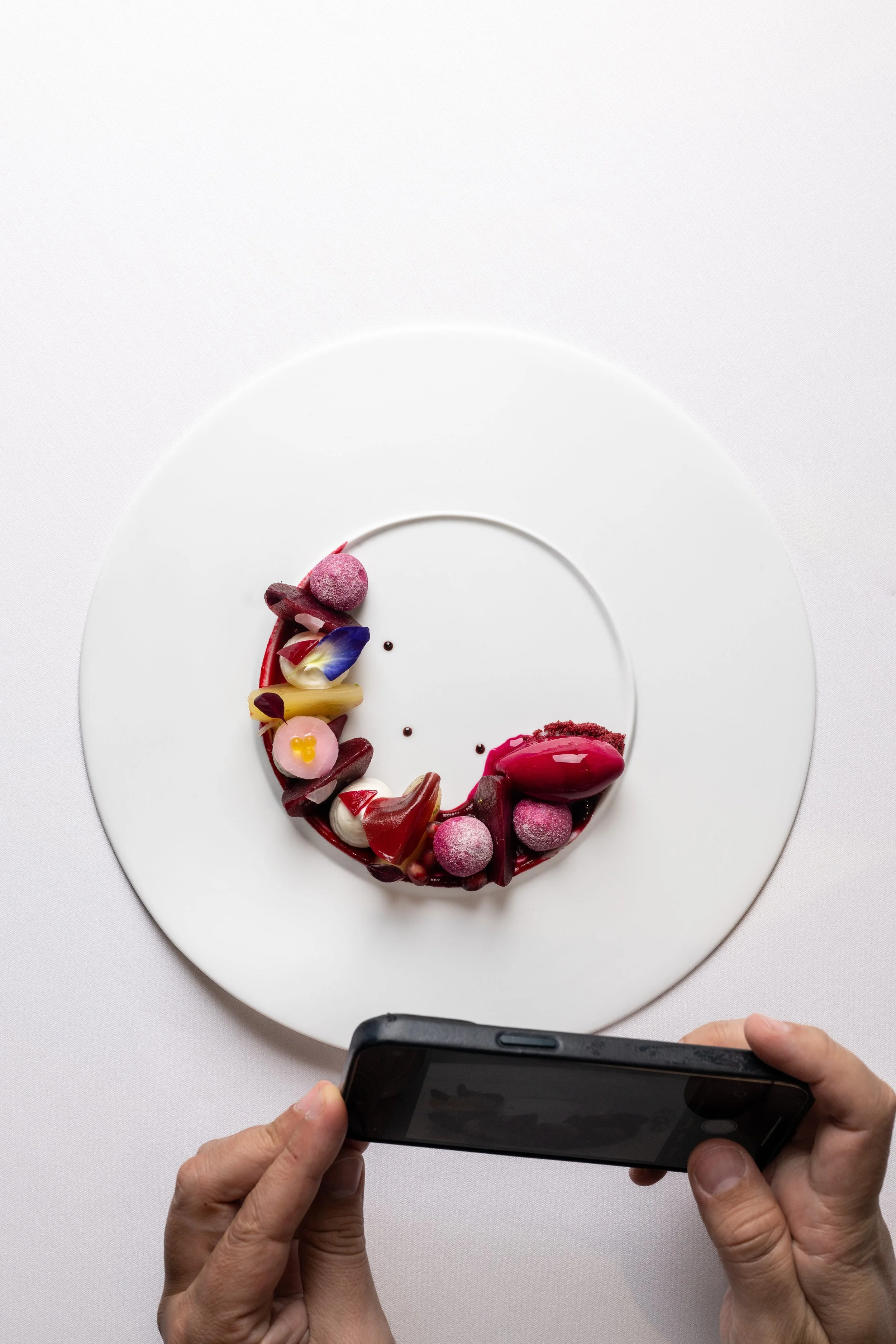 Plate with an artistic dessert arranged in the shape of a crescent moon, with various colorful garnishes and small decorative elements, and a person's hands holding a smartphone taking a photo of it.