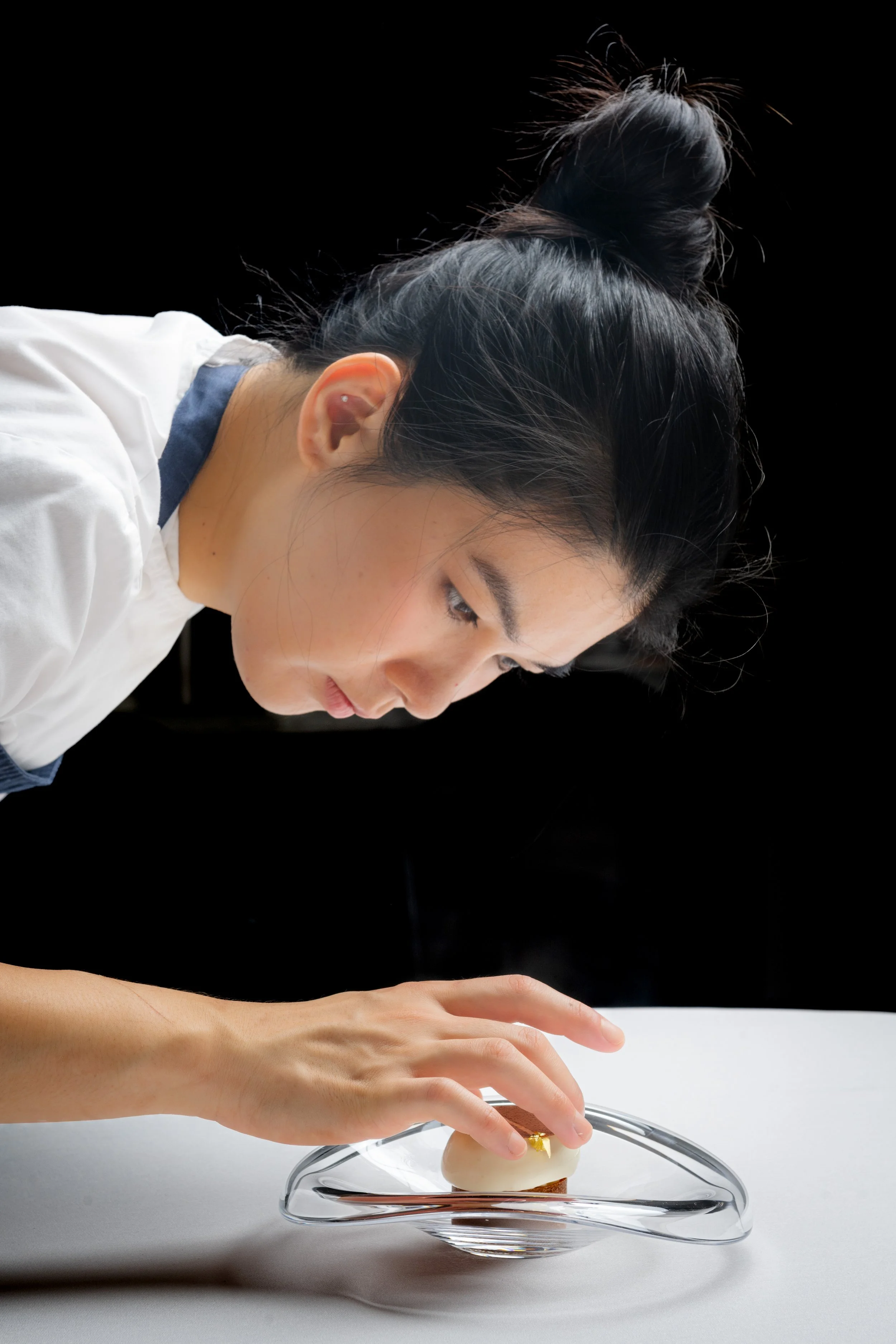 A woman with dark hair styled in a top bun, wearing a white shirt, is carefully placing a small item on a glass dish.