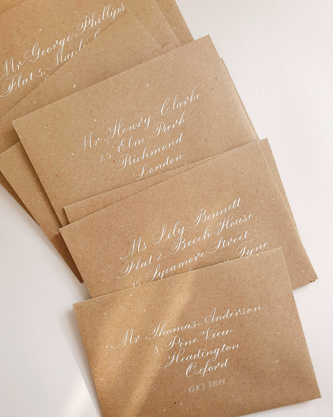 A close up of six kraft brown envelopes on a white background with addresses written on the front of each in copperplate calligraphy with white ink.