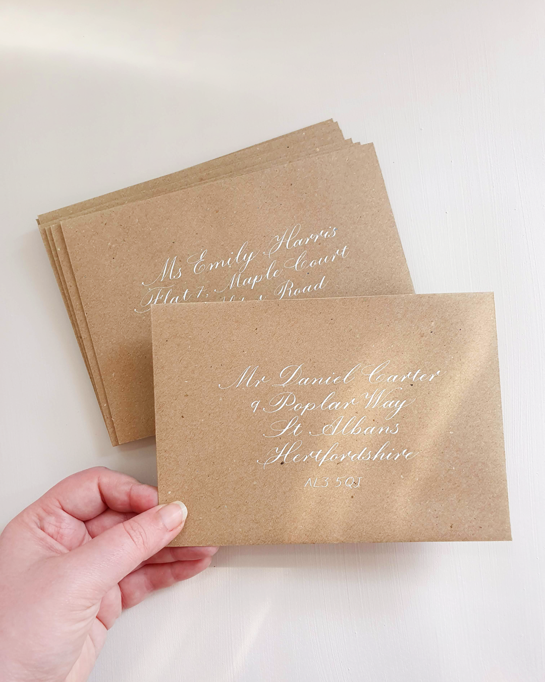 A hand is holding a kraft brown envelope against a white background with an address written on the front in copperplate calligraphy with white ink.